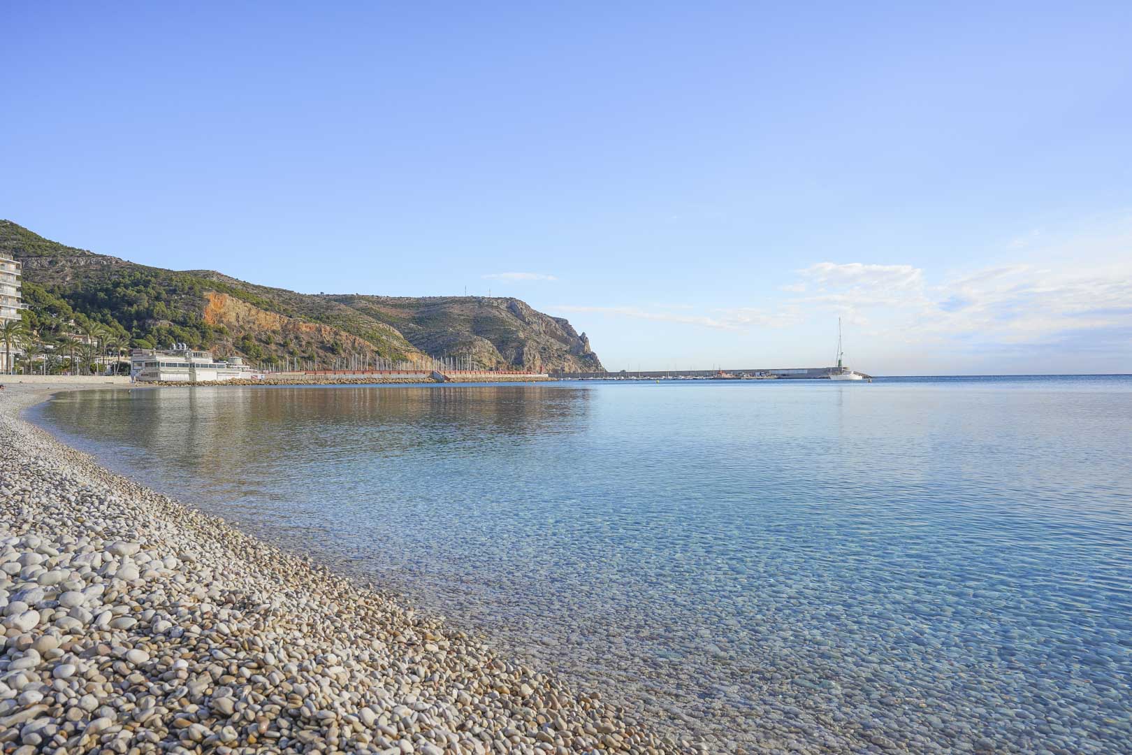 Panoramisch uitzicht op het kiezelstrand en het kristalheldere water in Jávea, met de haven en kustkliffen op de achtergrond onder een heldere hemel.