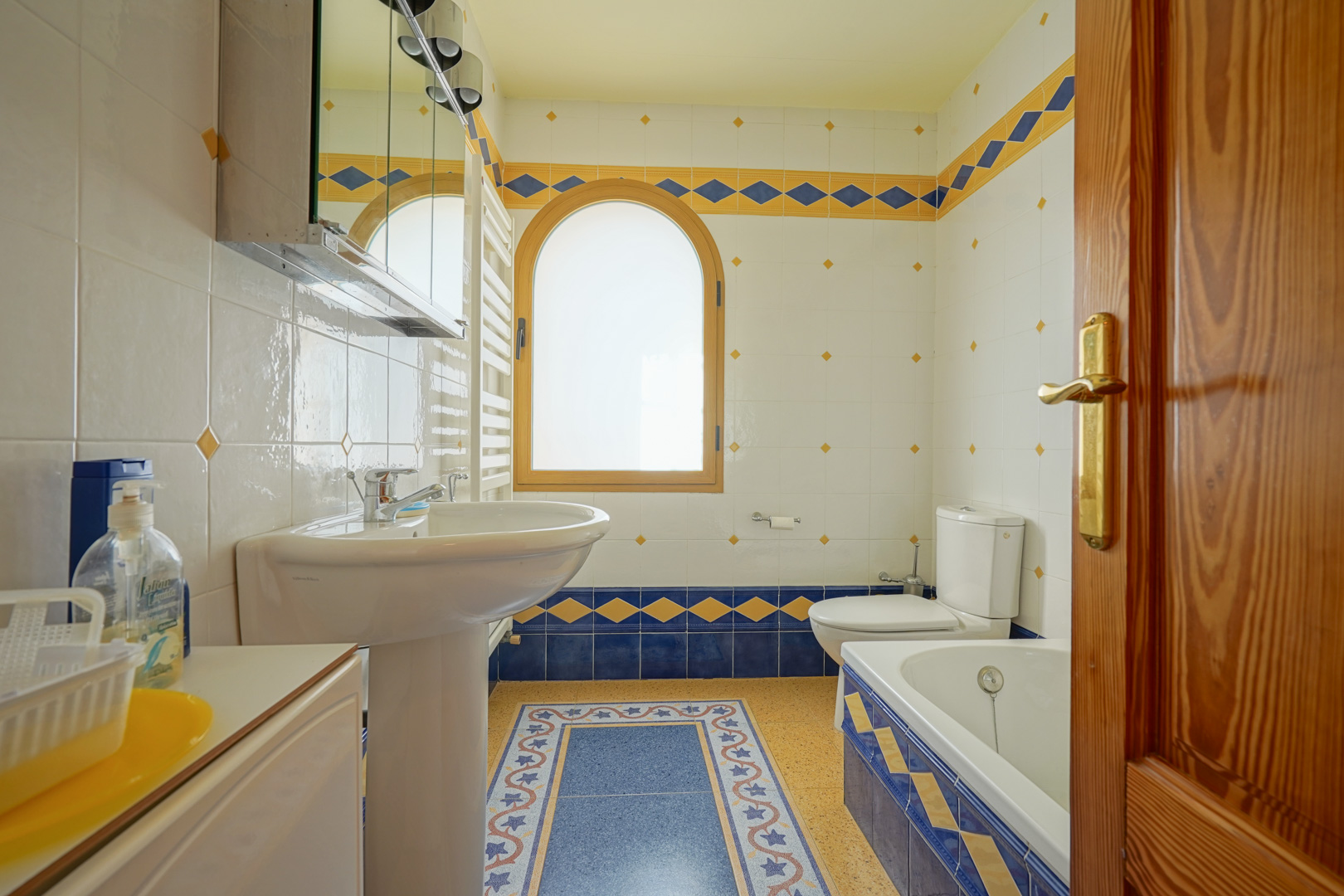 Badkamer met decoratieve blauwe en gele tegels, een rond houten boogvenster en een praktische indeling met ligbad, kolomwastafel en handdoekradiator.
