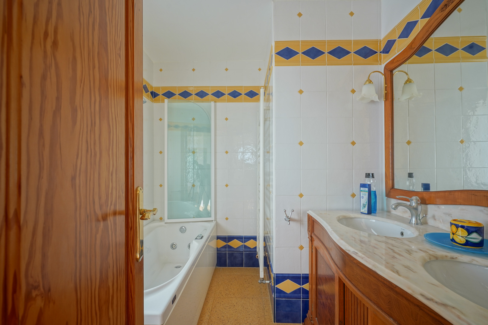 Badkamer met dubbele marmeren wastafel, houten meubel en hydromassagebad, afgewerkt met decoratieve blauwe en gele tegels in mediterrane stijl.