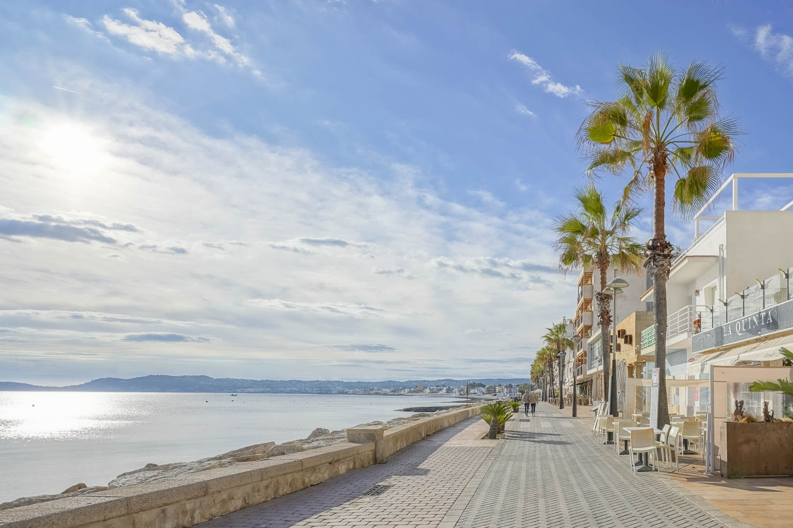 De boulevard van Jávea met palmbomen, terrassen en een stenen zeemuur, met weids uitzicht over de baai en de omliggende bergen in de zon.