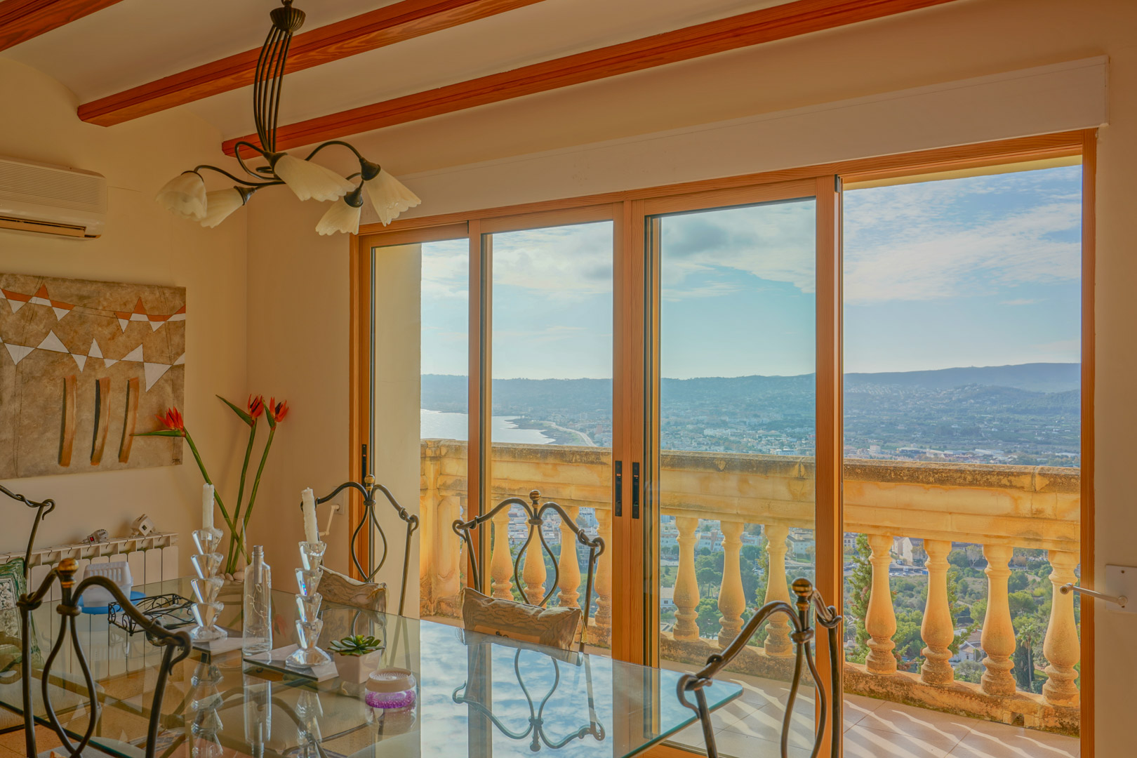 Eethoek met glazen tafel en houten plafondbalken, met grote schuifdeuren die een panoramisch uitzicht bieden op de baai van Jávea en de kust.