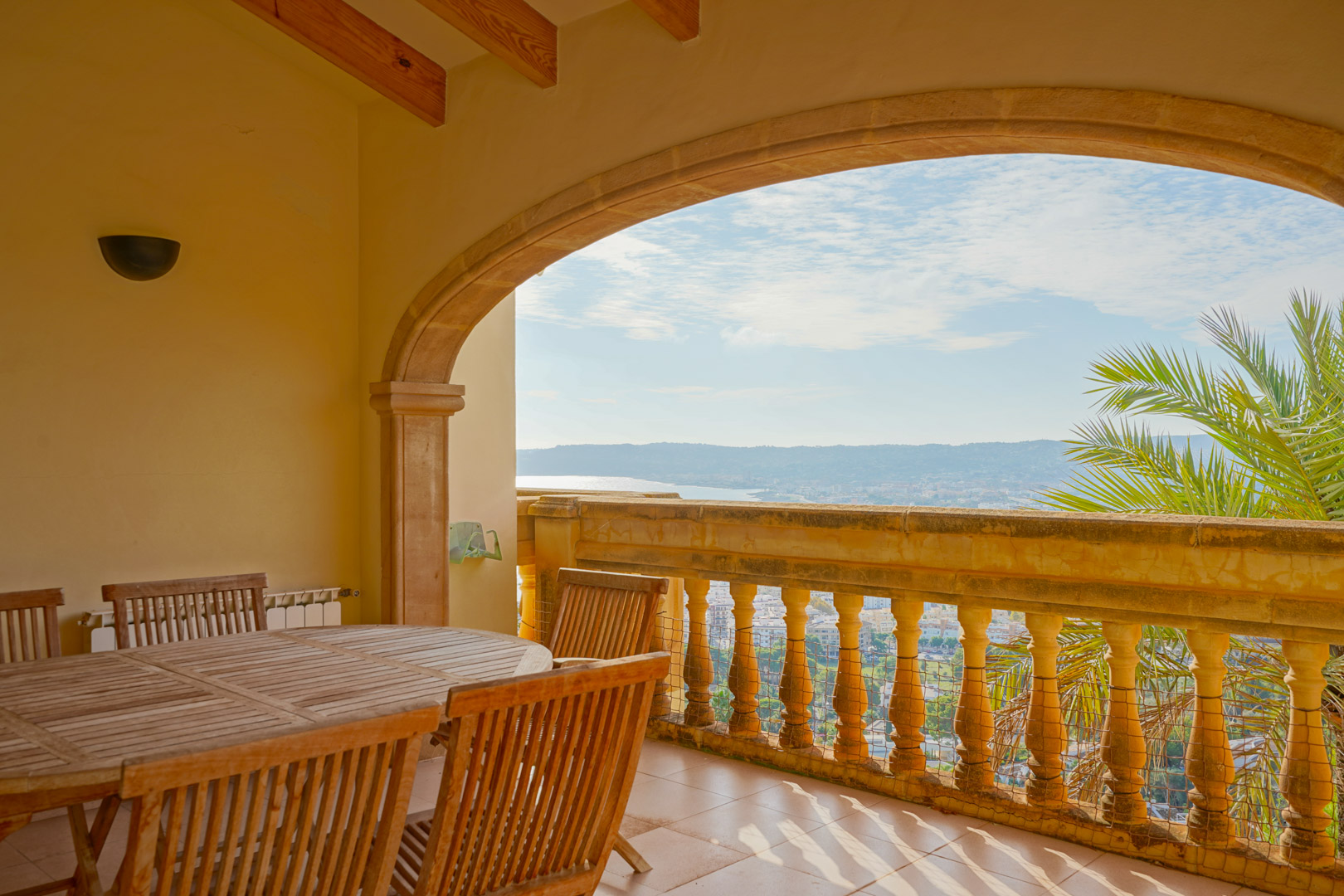 Overdekt terras met een boog van natuursteen, houten balkenplafond en een eethoek met panoramisch uitzicht over de baai van Jávea en de bergen.