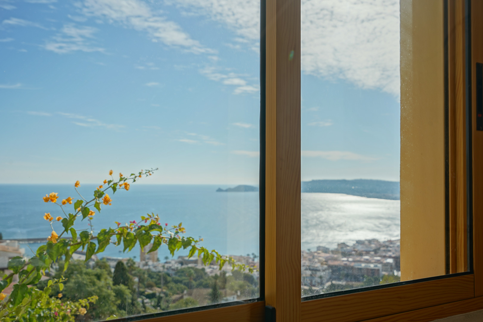 Panoramisch uitzicht over de baai van Jávea en Cap Negre door een houten raamkozijn, met bloeiende bougainville op de voorgrond.