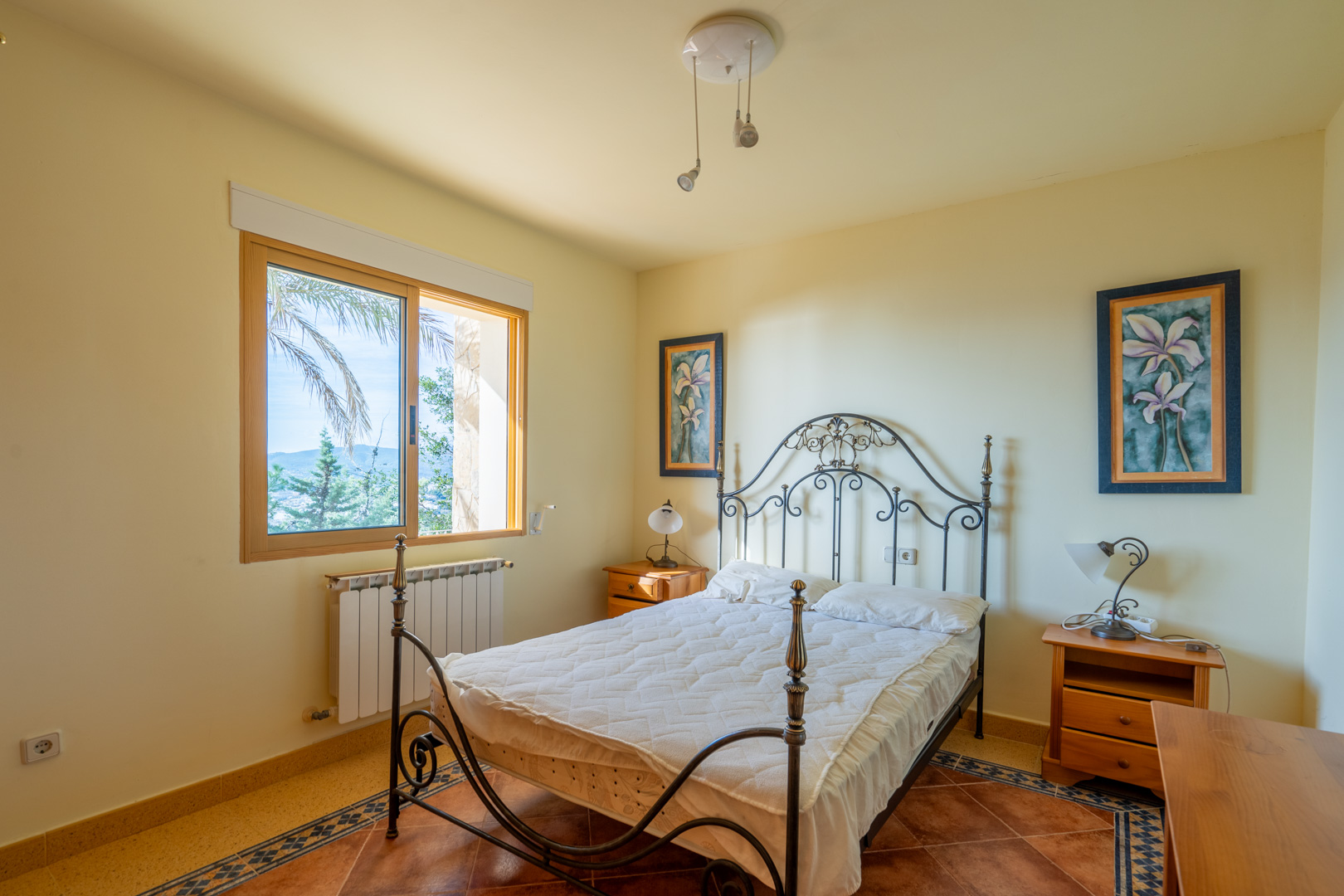 Slaapkamer met smeedijzeren bedframe, terracotta tegelvloer met decoratieve rand en houten raam met uitzicht op de omliggende natuur.