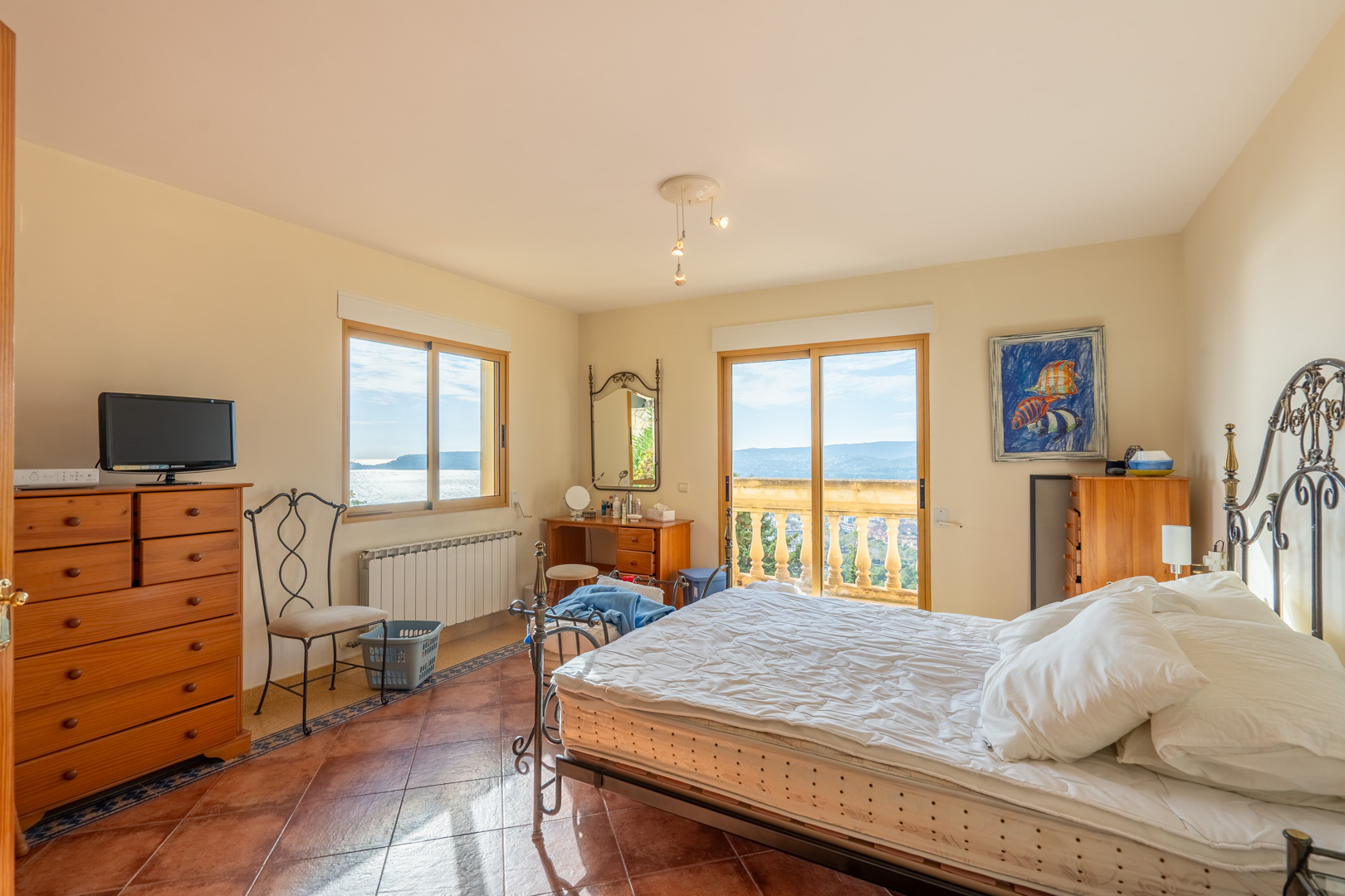 Slaapkamer met terracotta vloertegels, een smeedijzeren bed en schuifdeuren naar een balkon met stenen balustrades en panoramisch uitzicht over de baai.