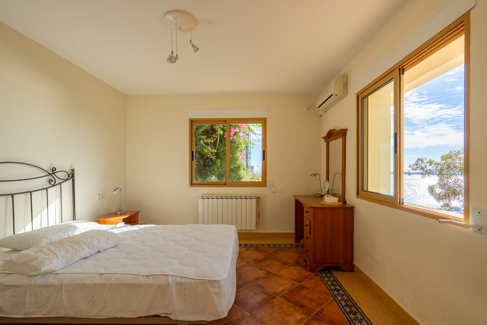 Slaapkamer met terracotta vloertegels en decoratieve randen. Voorzien van houten kozijnen met zeezicht, een smeedijzeren hoofdbord en airconditioning.