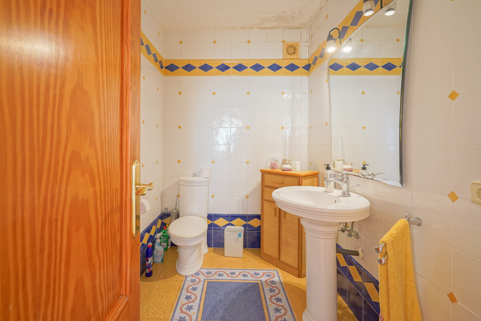 Traditionele badkamer met witte wandtegels, blauw-gele ruitpatronen, een klassieke kolomwastafel en een houten opbergkastje.