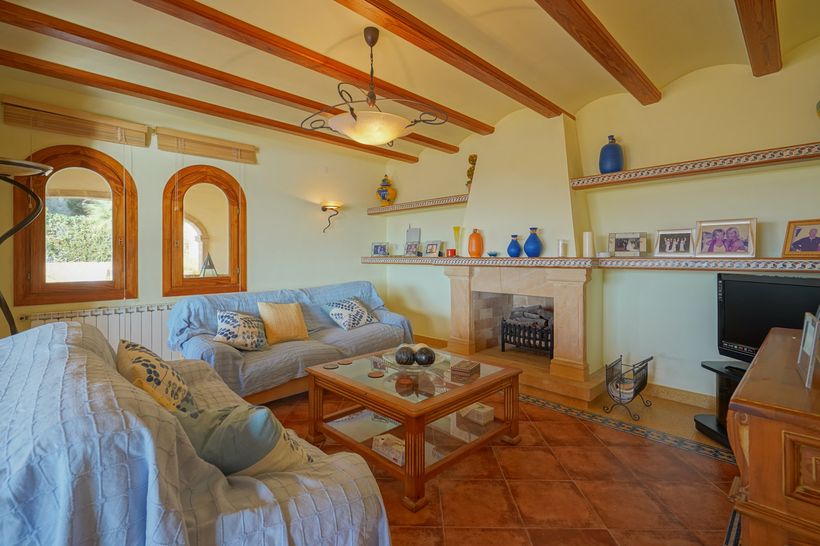 Traditionele woonkamer met stenen open haard, houten plafondbalken en terracotta vloertegels. Voorzien van houten boogramen en ingebouwde planken met keramische details.
