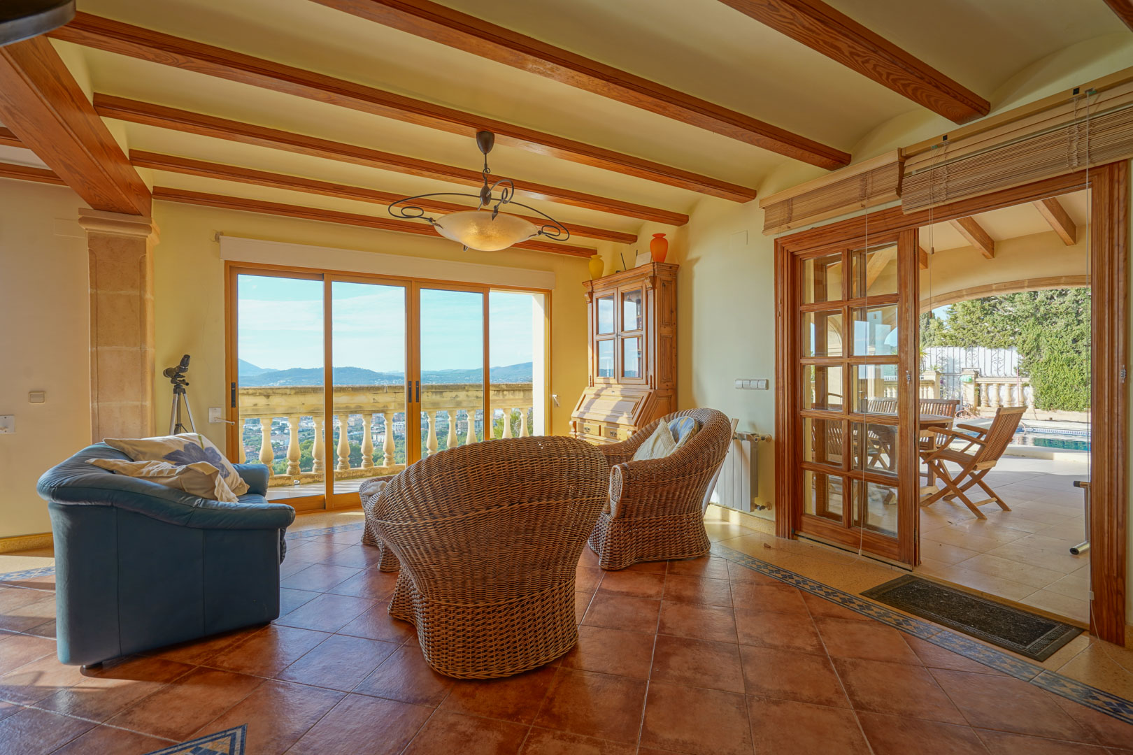 Woonkamer met houten balkenplafond, terracotta vloer en rotan stoelen. Grote glazen deuren bieden panoramisch uitzicht op de vallei en toegang tot het terras bij het zwembad.