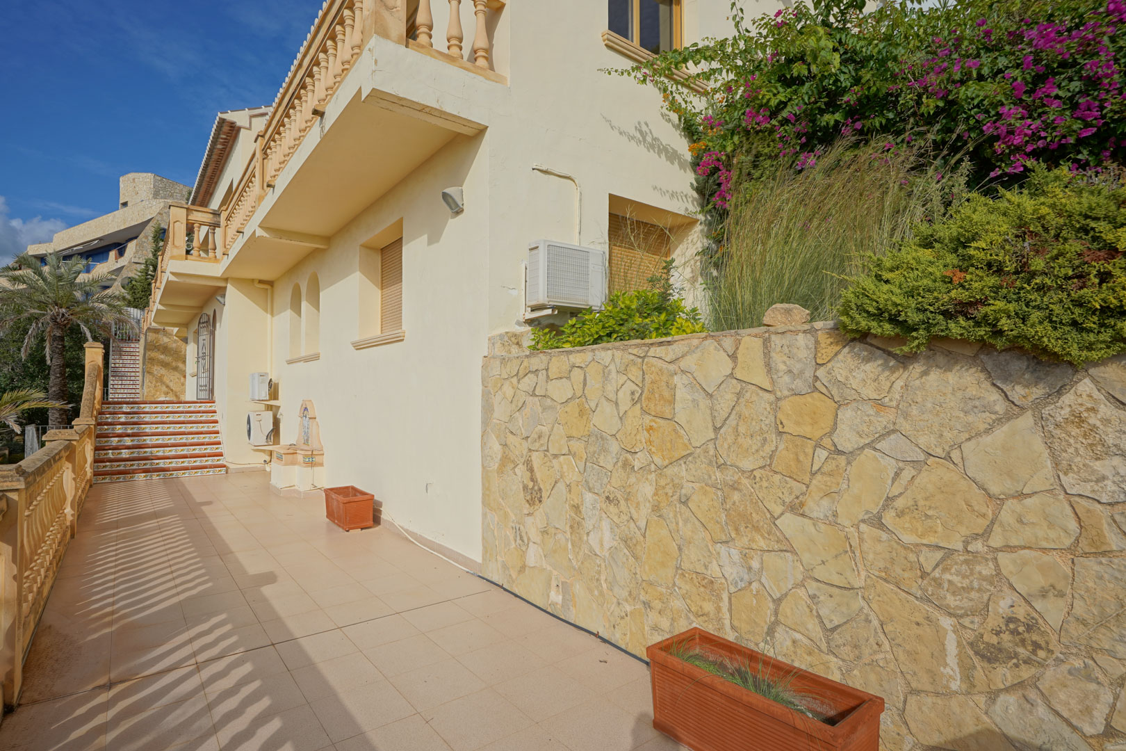 Zijterras met natuurstenen muur, terracotta plantenbakken en trappen met traditionele decoratieve tegels. De gevel toont klassieke mediterrane architectonische details.