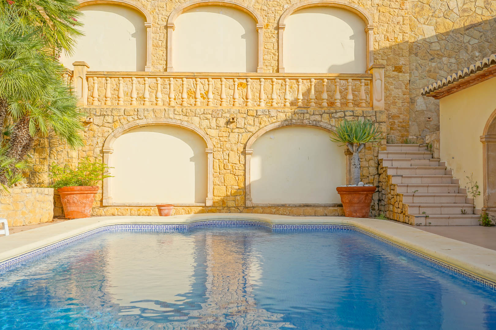 Zwembad met blauwe mozaïektegels en een klassieke bies, geflankeerd door natuurstenen muren met boognissen, een stenen balustrade en terracotta plantenbakken.