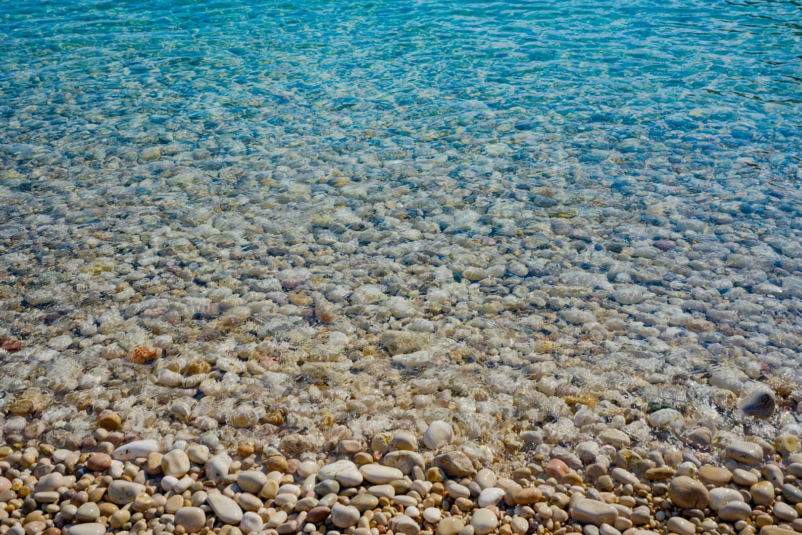 Gedetailleerd zicht op de kustlijn van het Granadella-strand, met gladde witte kiezelstenen en kristalhelder turquoise mediterraan water.