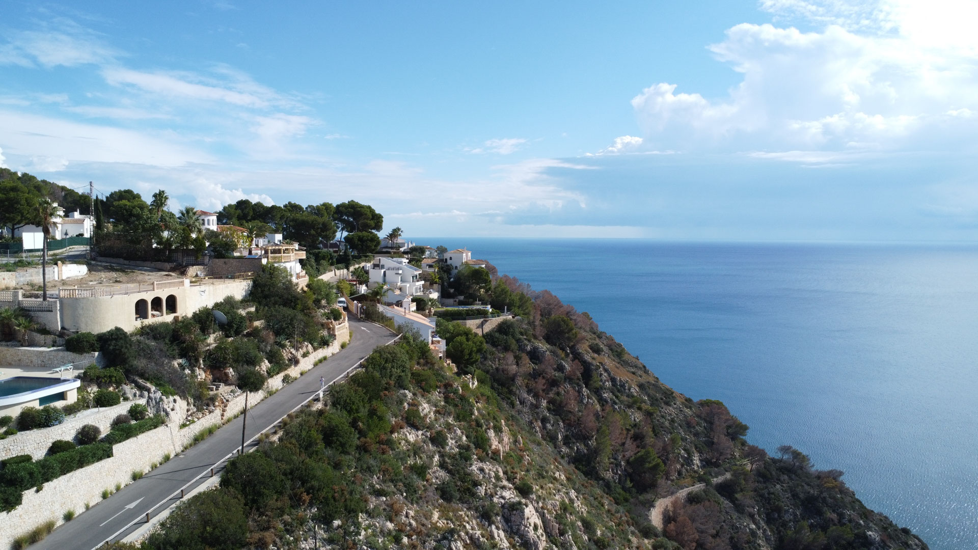 Luchtfoto van de kustlijn van Granadella in Jávea, met percelen op een helling, direct uitzicht op de Middellandse Zee en een geasfalteerde toegangsweg.