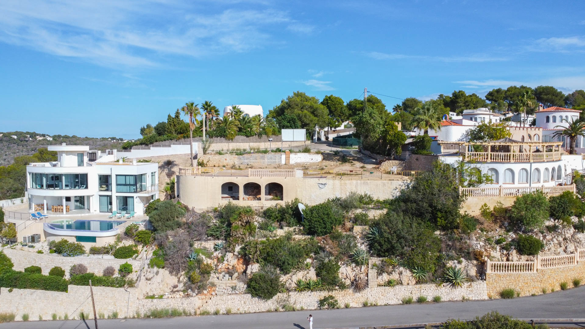 Luchtfoto van het hooggelegen perceel met een boogvormige stenen fundering, gelegen tussen luxe villa's en mediterrane beplanting op stenen muren.