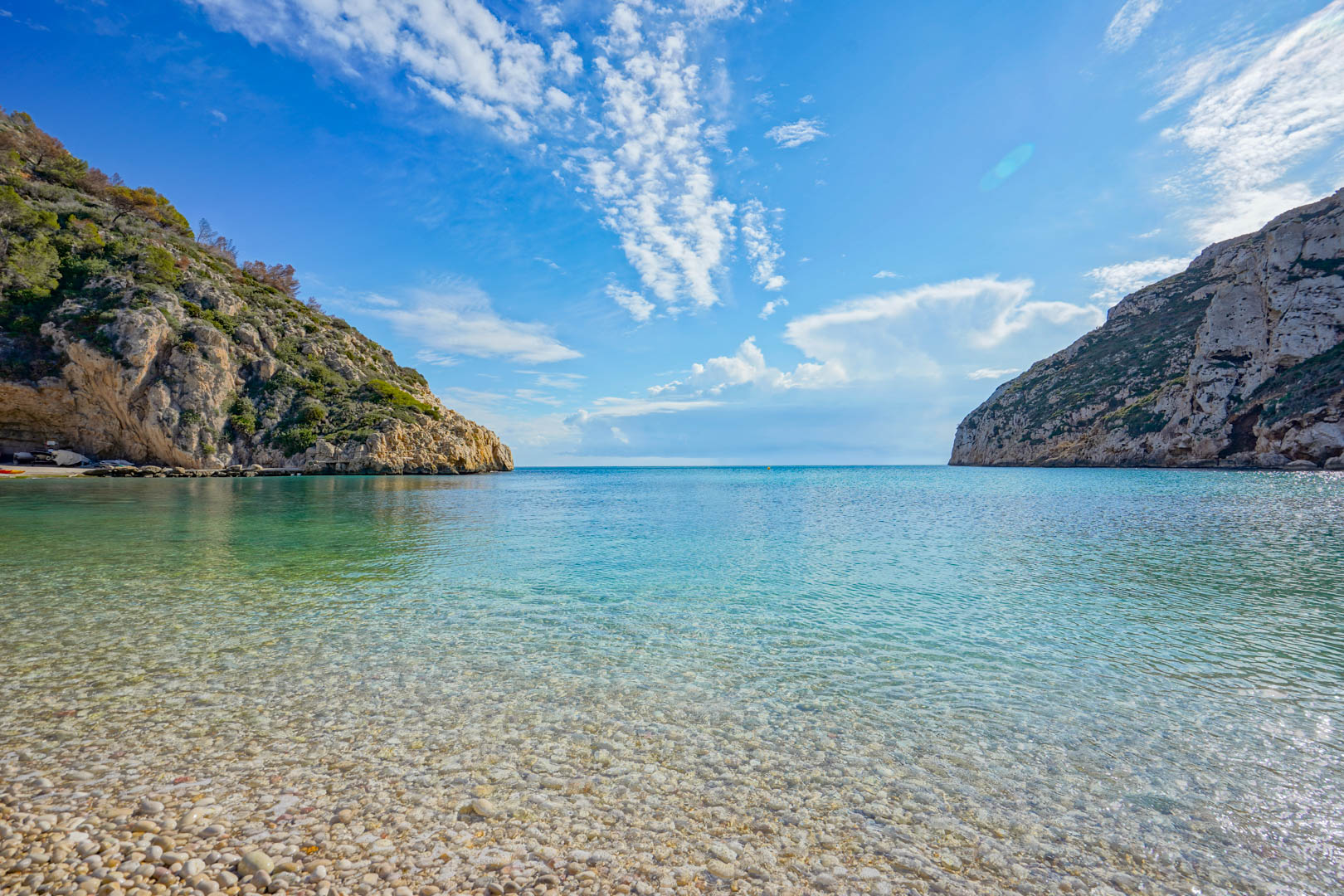 Panoramisch uitzicht op het strand van Granadella met kristalhelder turquoise water en een kiezelstrand, omgeven door steile mediterrane kliffen en een helderblauwe lucht.