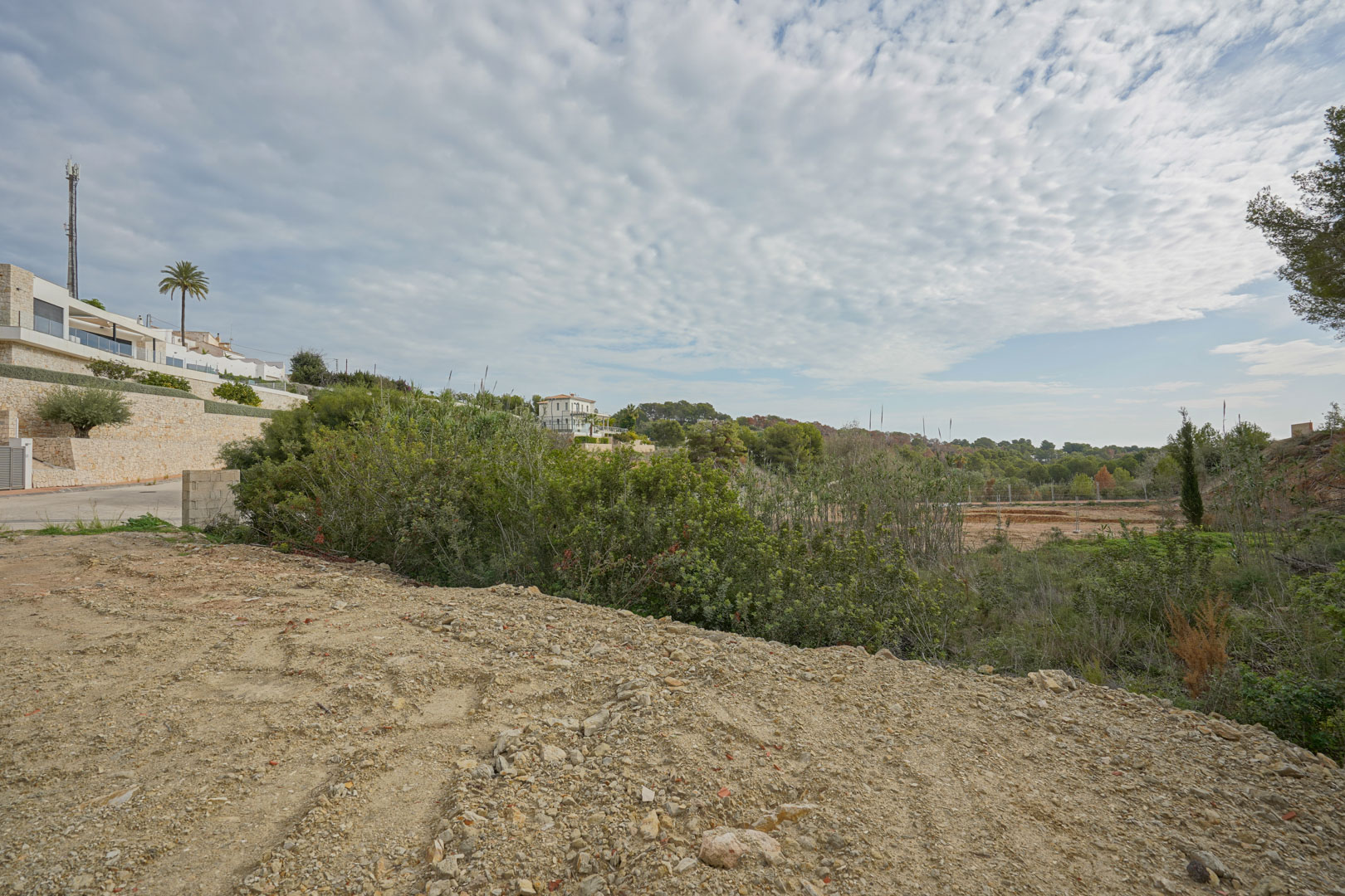 Bouwrijp perceel omgeven door mediterrane vegetatie en moderne villa's met natuurstenen muren in een rustige woonwijk.