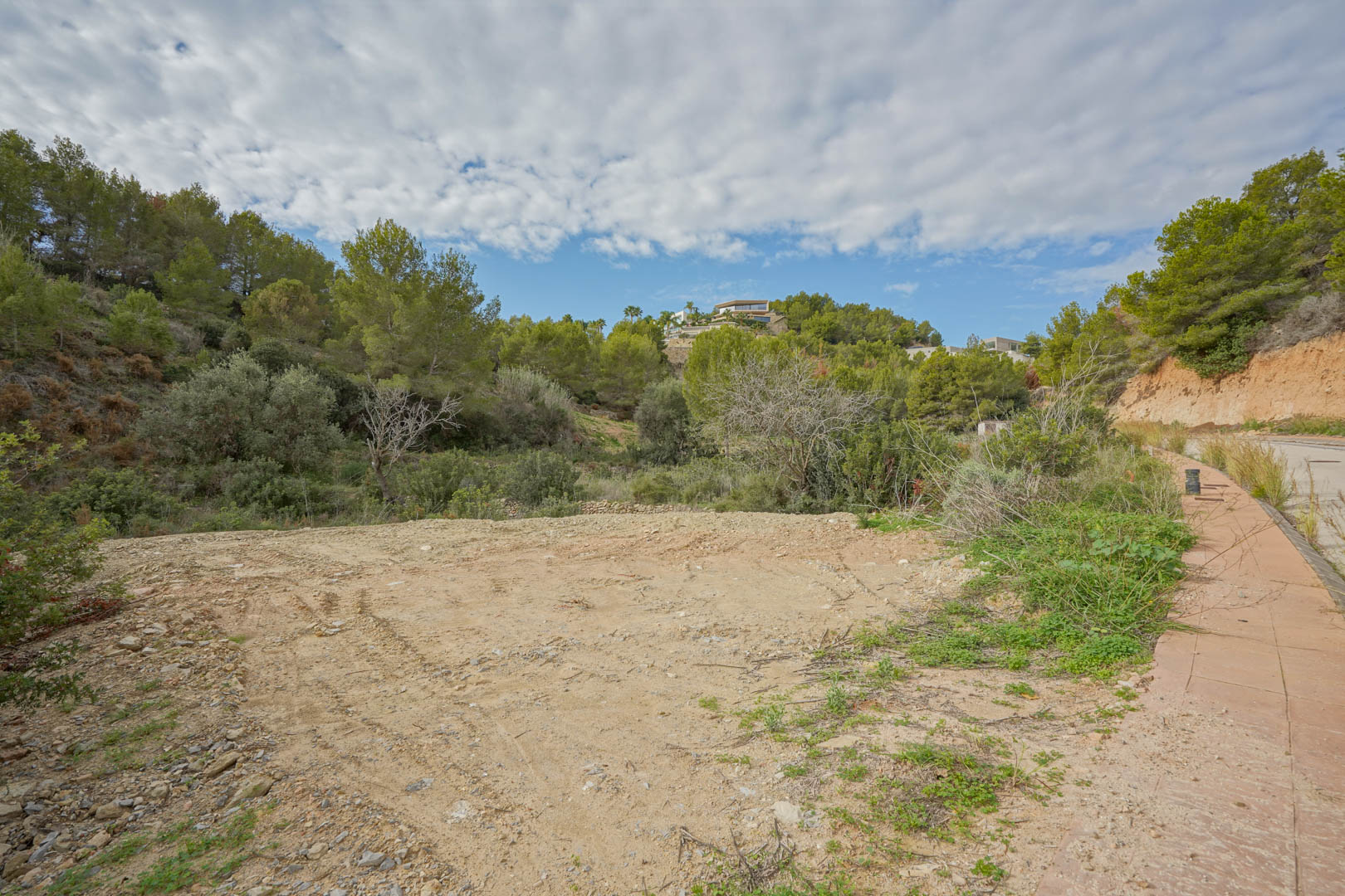 Zicht op het vlakke, bouwrijpe perceel omgeven door mediterrane pijnbomen, met directe toegang vanaf een verharde weg in een moderne woonwijk.