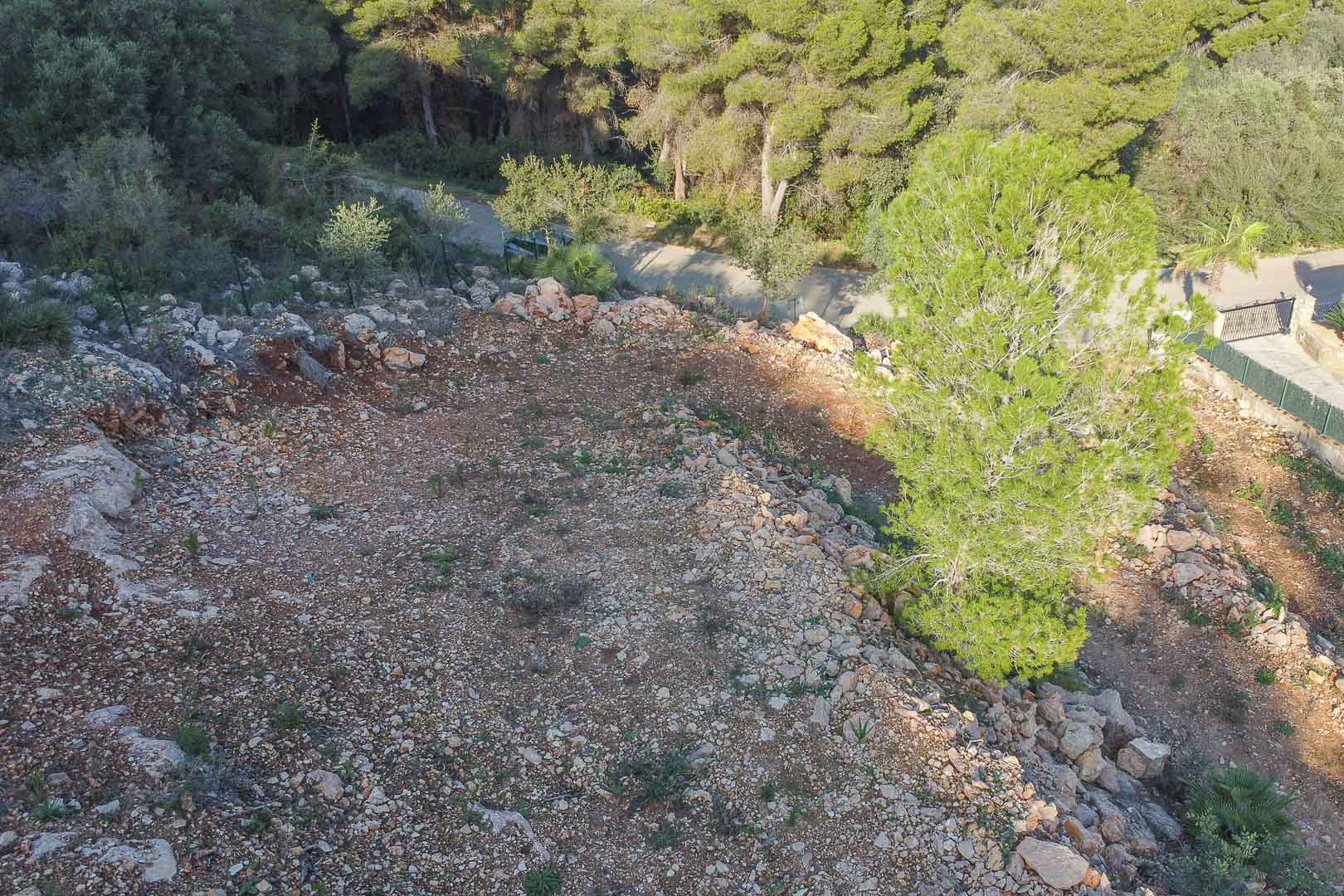 Verhoogd zicht op het bouwperceel met natuurlijke topografie van rode aarde en rotsen, omgeven door een dicht mediterraan dennenbos.
