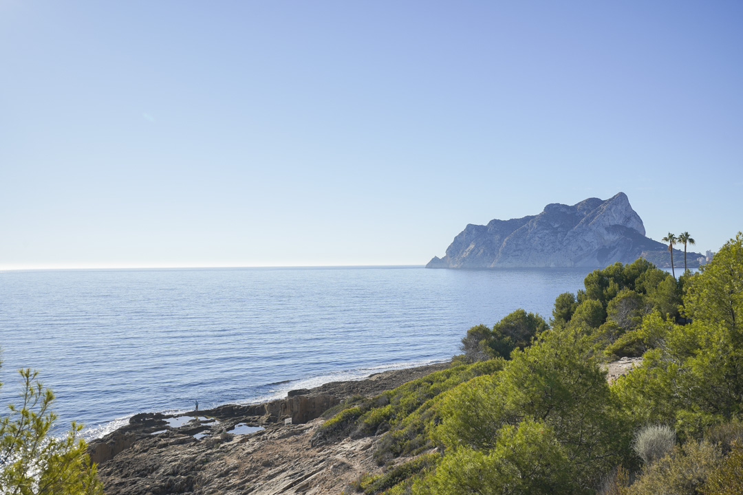 Panoramisch uitzicht op de Peñón de Ifach en de Middellandse Zeekust, met rotsachtige oevers en inheemse pijnbomen onder een heldere lucht.