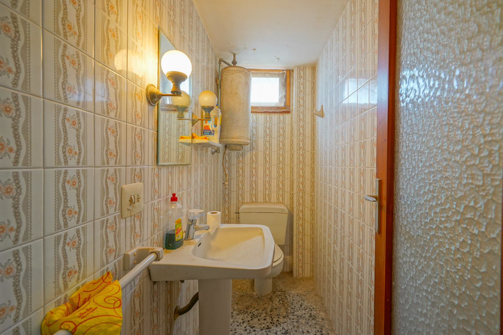 Functionele badkamer met vintage wandtegels met patroon, een wastafel op zuil, elektrische boiler en originele terrazzovloer.