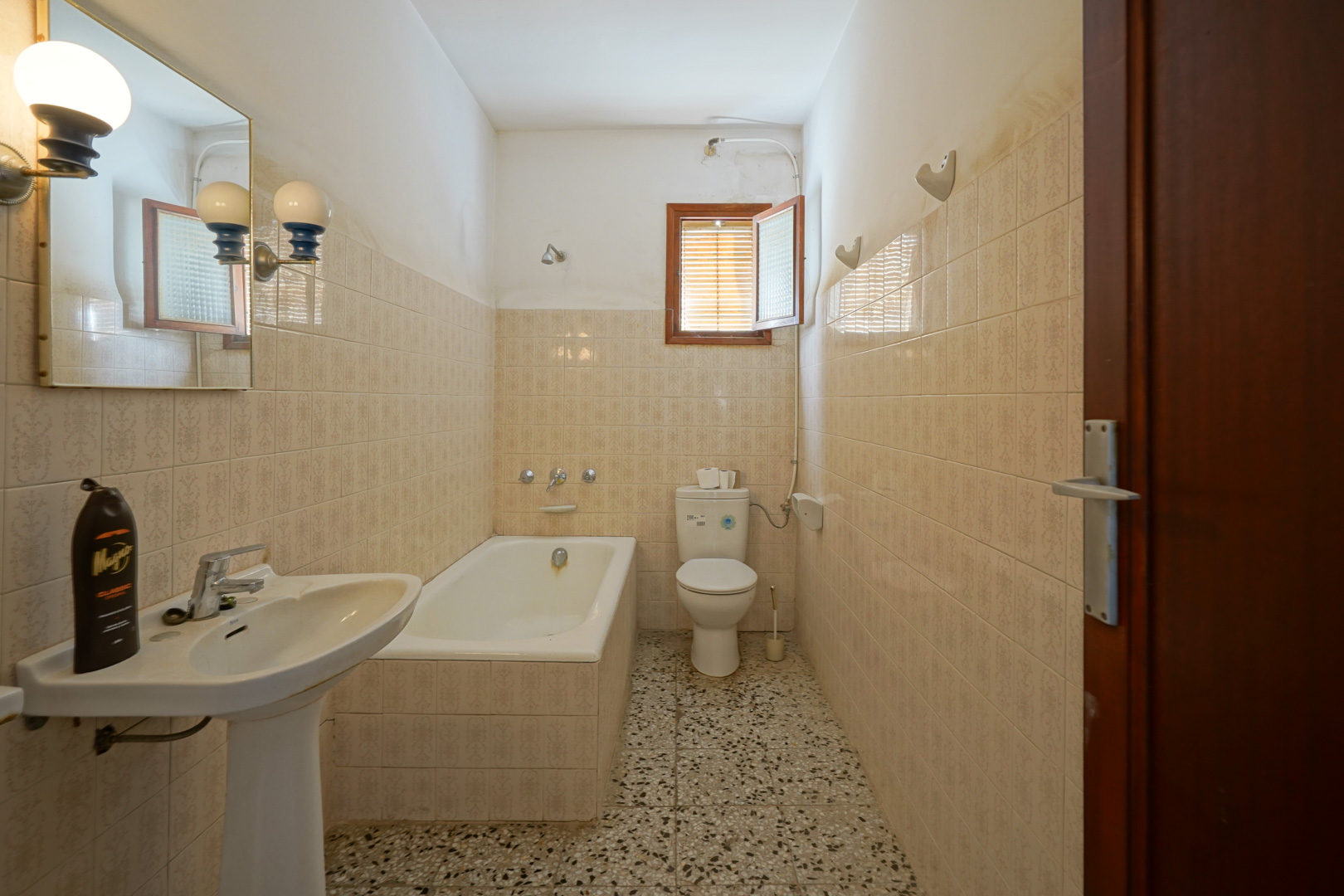 Functionele badkamer voorzien van een ingebouwd ligbad, kolomwastafel, terrazzovloer en klassiek betegelde wanden met een klein raam.