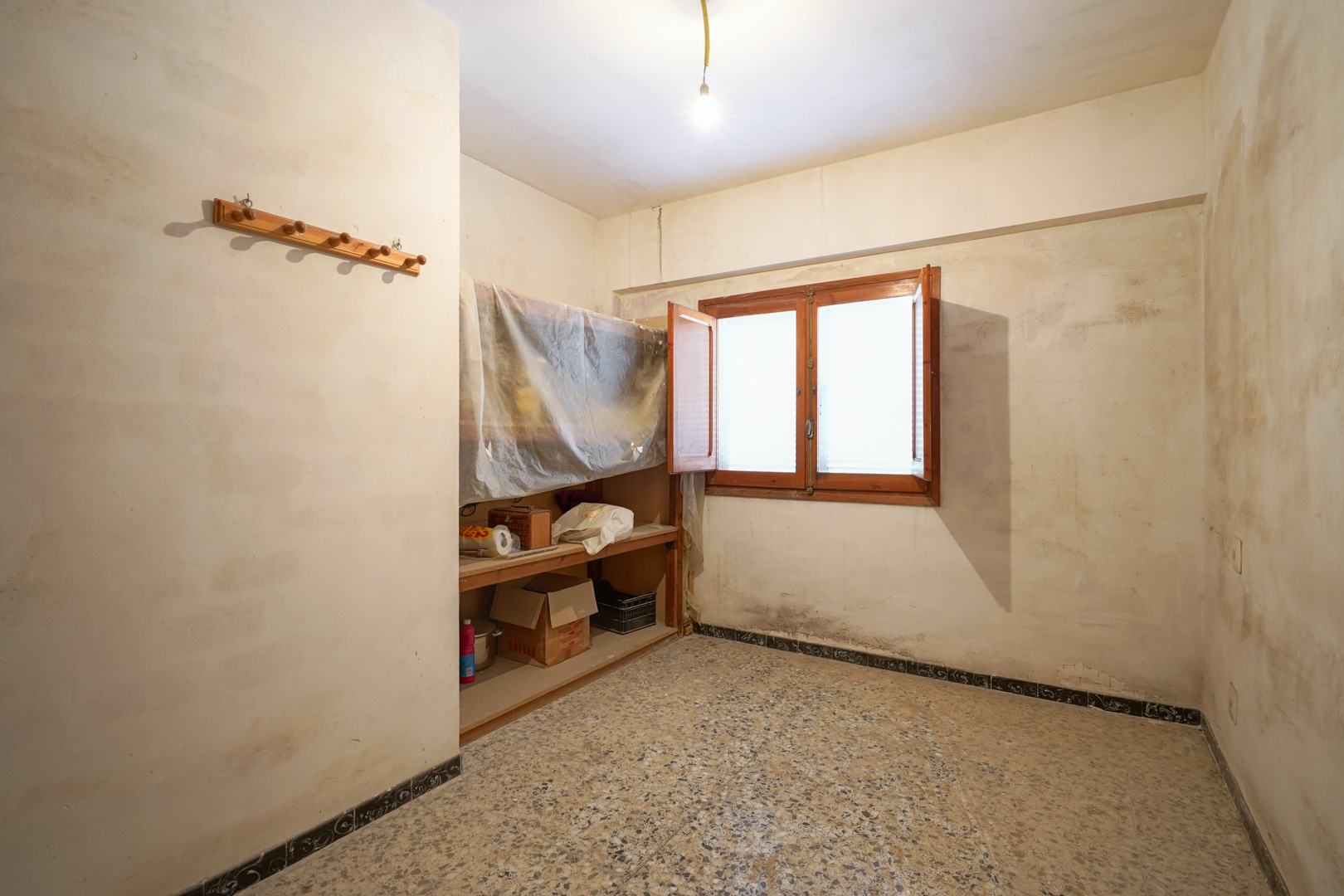 Kamer met terrazzo vloer, houten venster met dubbele vleugels en ingebouwde schappen, klaar voor een volledige renovatie.