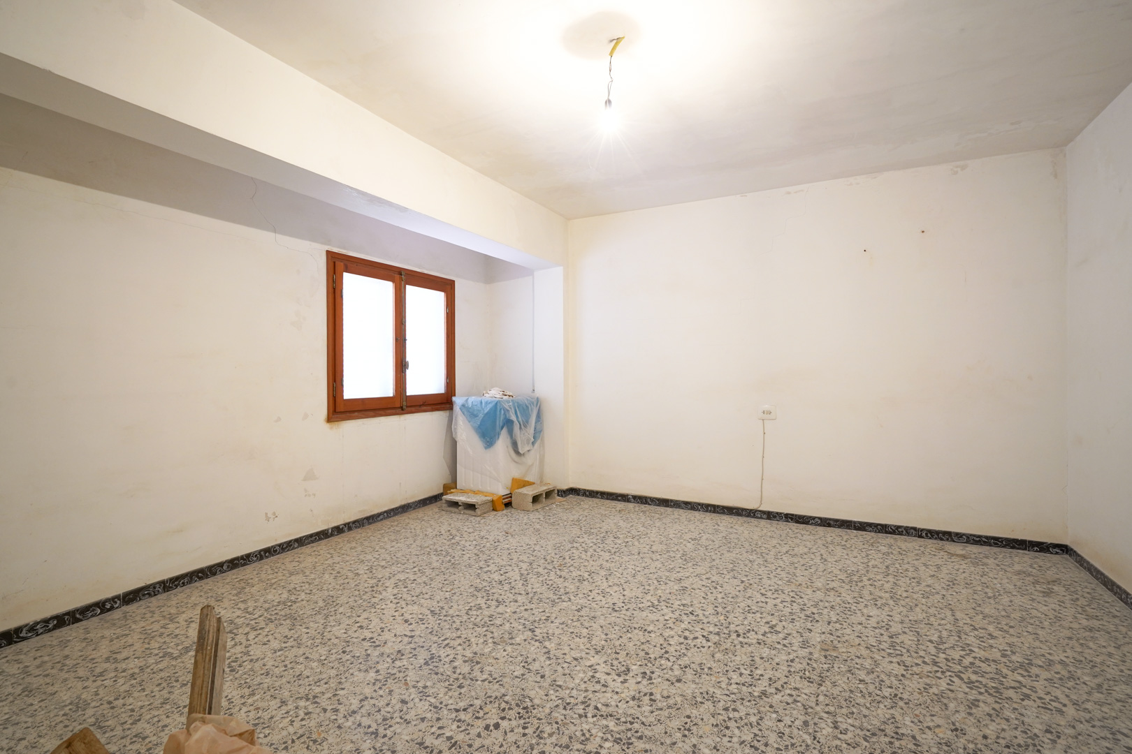 Ruime kamer met originele terrazzovloer, witte muren die renovatie behoeven en een houten raamkozijn voor natuurlijk licht.