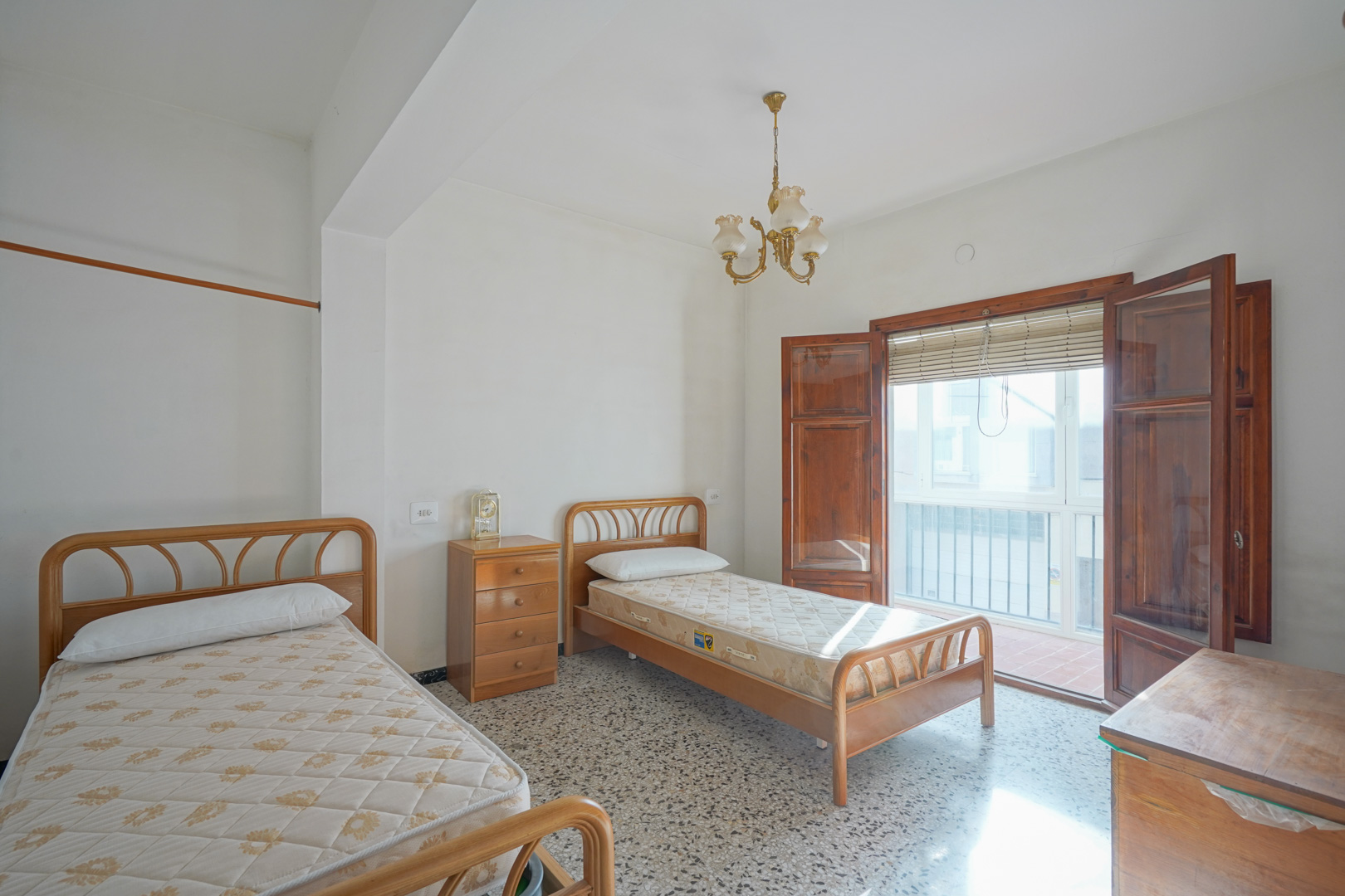 Slaapkamer met twee eenpersoonsbedden, terrazzo vloer en toegang tot een balkon via authentieke houten openslaande deuren.