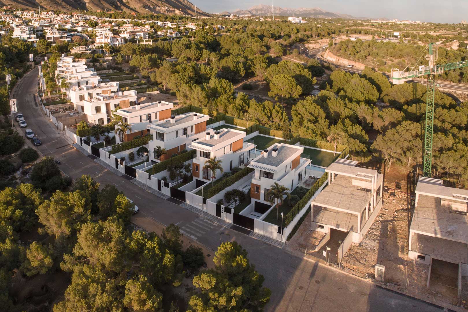 Luchtfoto van het wooncomplex met moderne witte villa's, houtaccenten en privézwembaden, gelegen in een natuurlijke omgeving van pijnbomen en bergen.