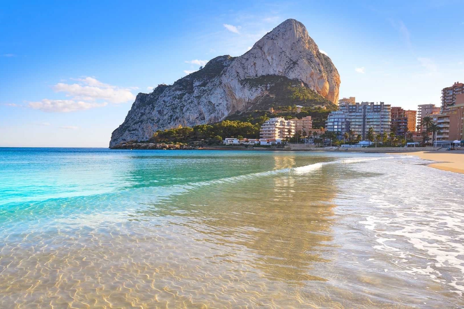 Panoramisch uitzicht op de Peñón de Ifach vanaf de kustlijn van Calpe, met kristalhelder turquoise water en de skyline van de kustgebouwen.