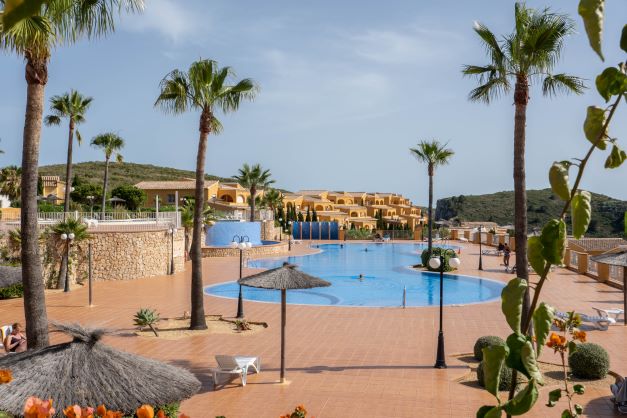 Groot gemeenschappelijk zwembad omgeven door een ruim terras van terracotta, palmbomen en rieten parasols in de urbanisatie Monte Cala Gardens.