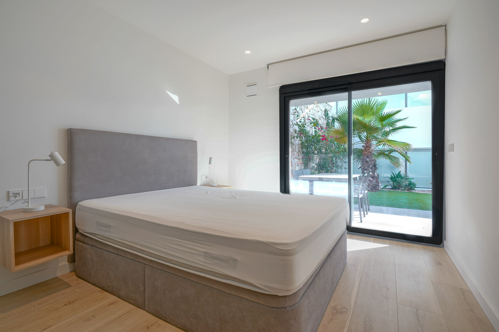 Hoofdslaapkamer met directe toegang tot de tuin via schuifdeuren, voorzien van een lichte houten vloer en een modern grijs gestoffeerd bed.