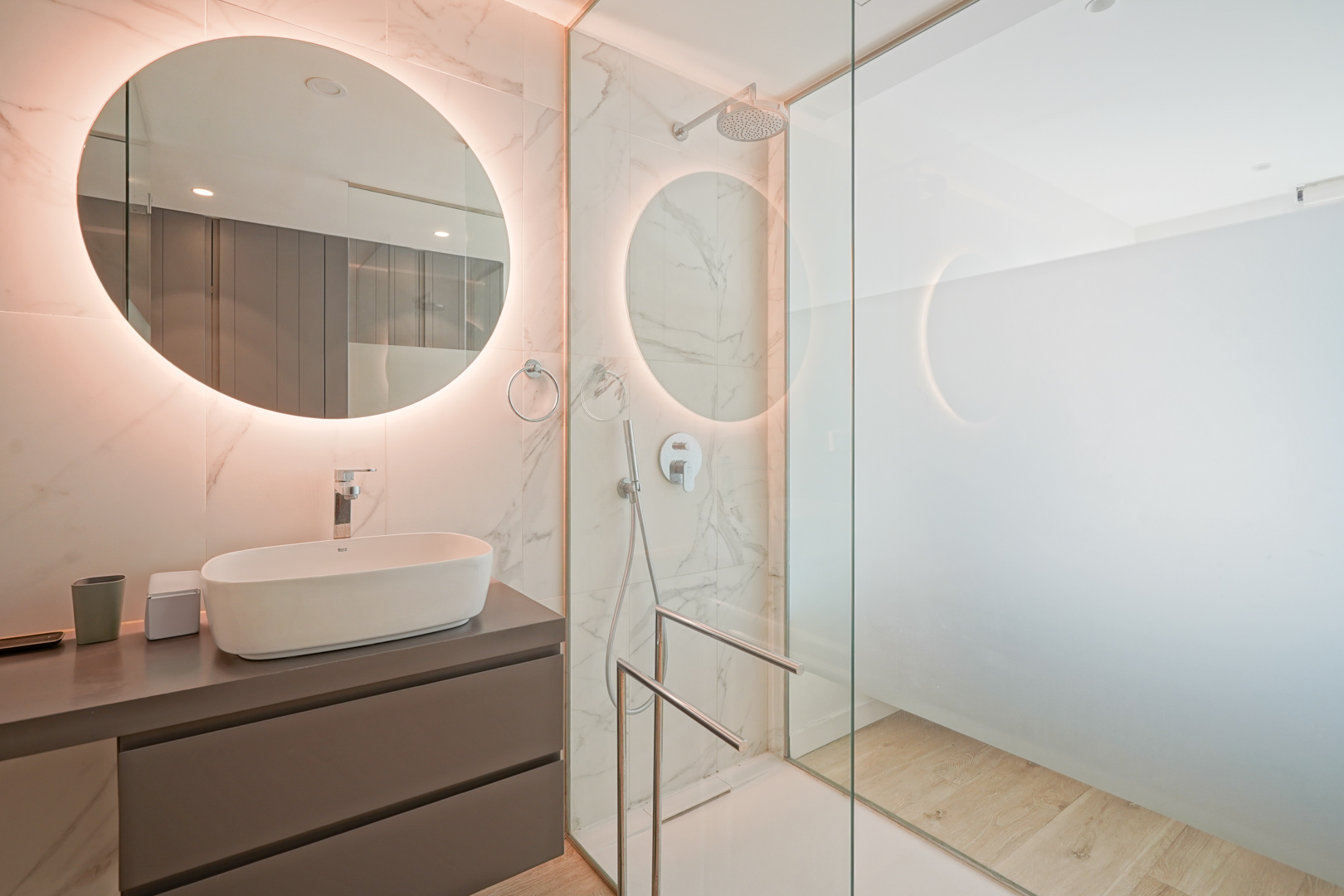 Moderne badkamer met opzetwastafel, verlichte ronde spiegel en inloopdouche met glazen wand en wandtegels met marmerlook.
