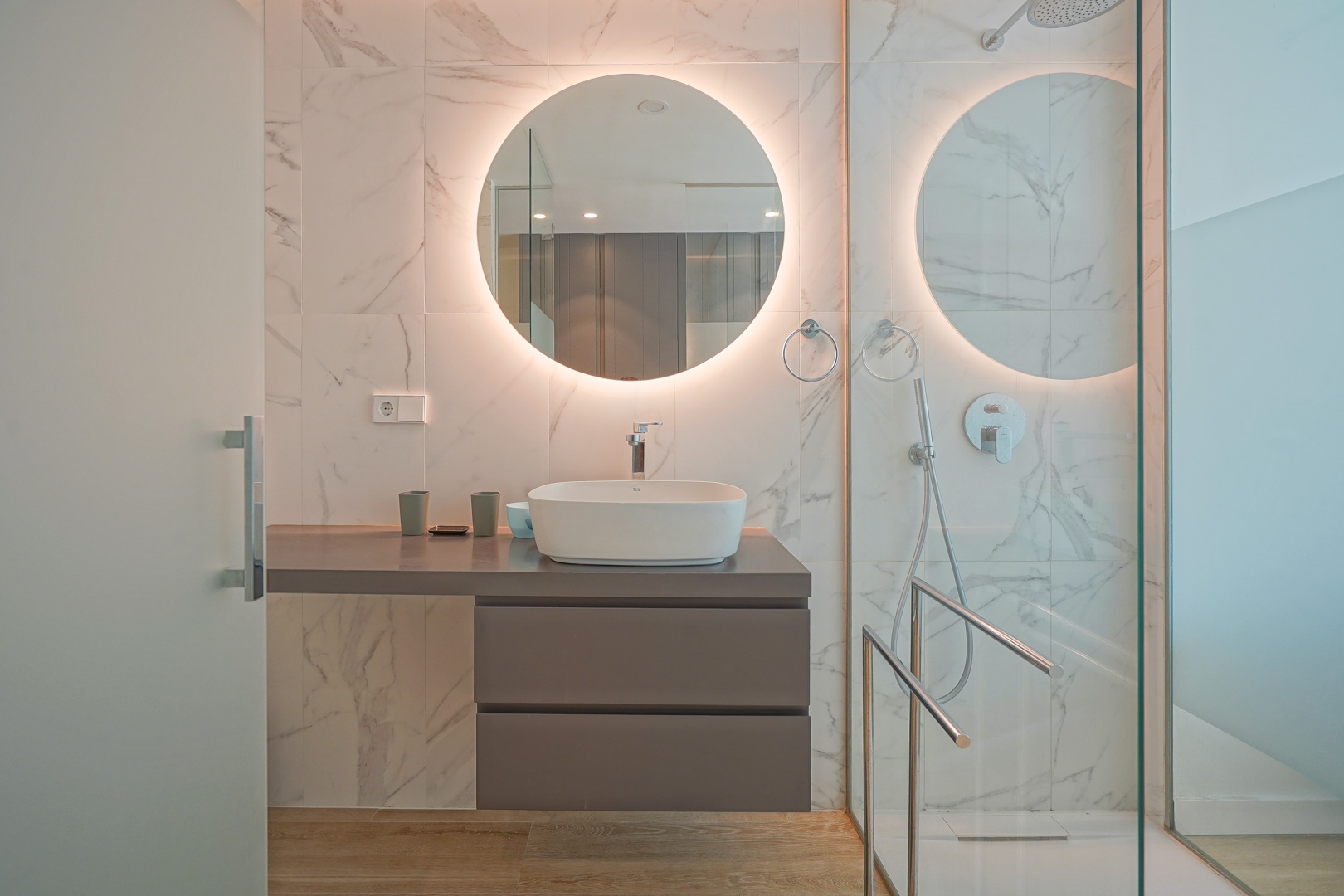 Moderne badkamer met opzetwastafel, verlichte ronde spiegel en wandtegels met marmerlook naast een glazen inloopdouche.