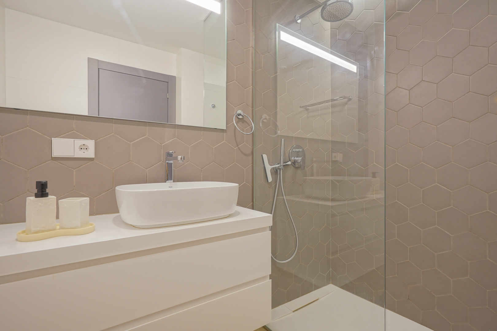 Moderne badkamer met taupekleurige zeshoekige tegels, een opzetwastafel op een wit meubel en een inloopdouche met glazen wand.
