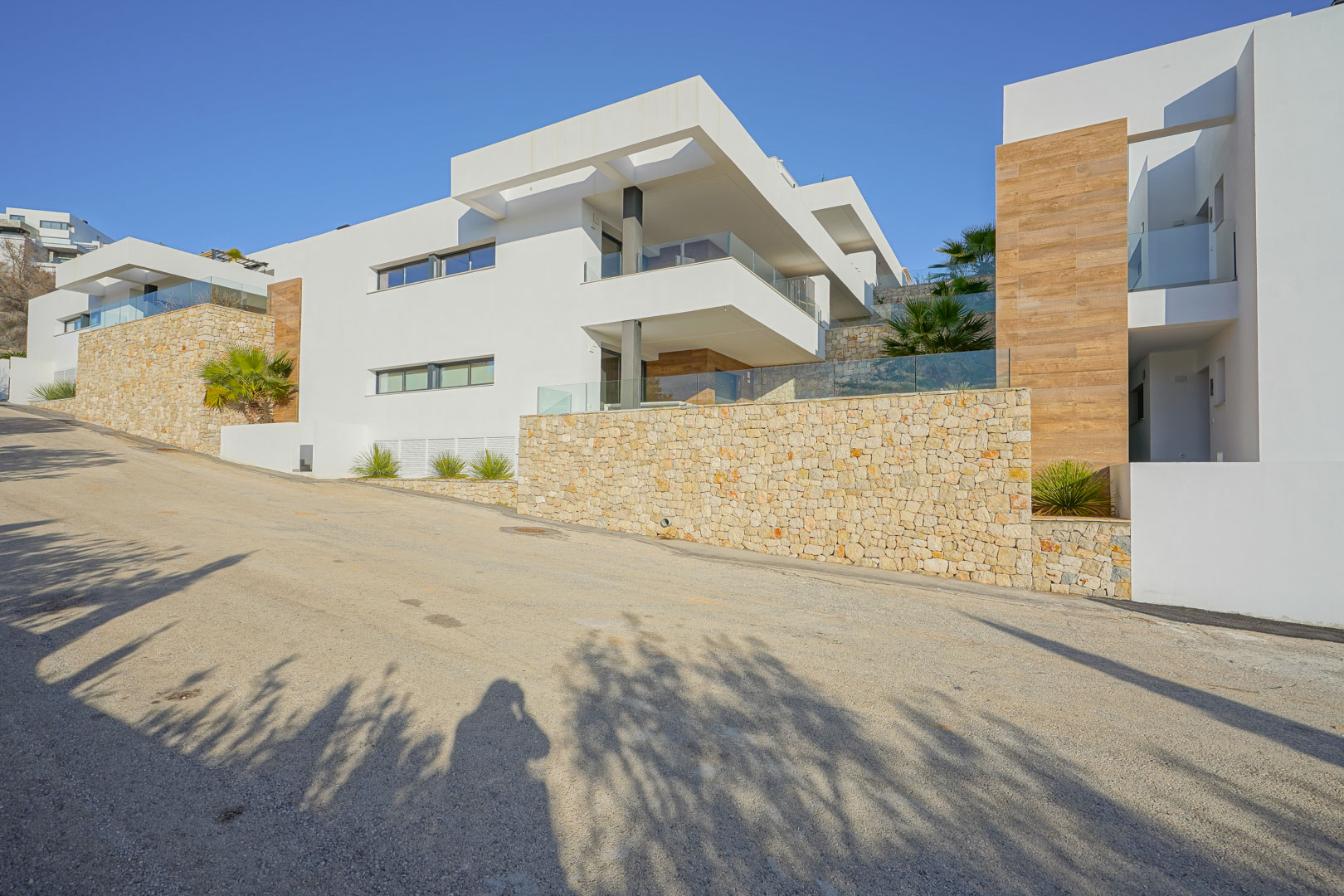Moderne gevel in Monte Cala Gardens met natuurstenen muren, ruime terrassen met glazen balustrades en houtaccenten tegen een helderblauwe lucht.