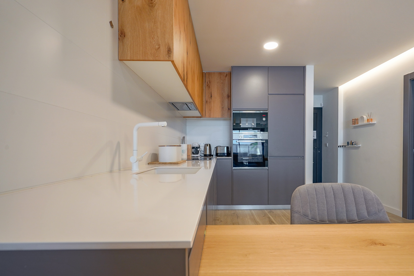 Moderne keuken met wit werkblad, grijze en houten kasten, geïntegreerde apparatuur en een minimalistische witte kraan.
