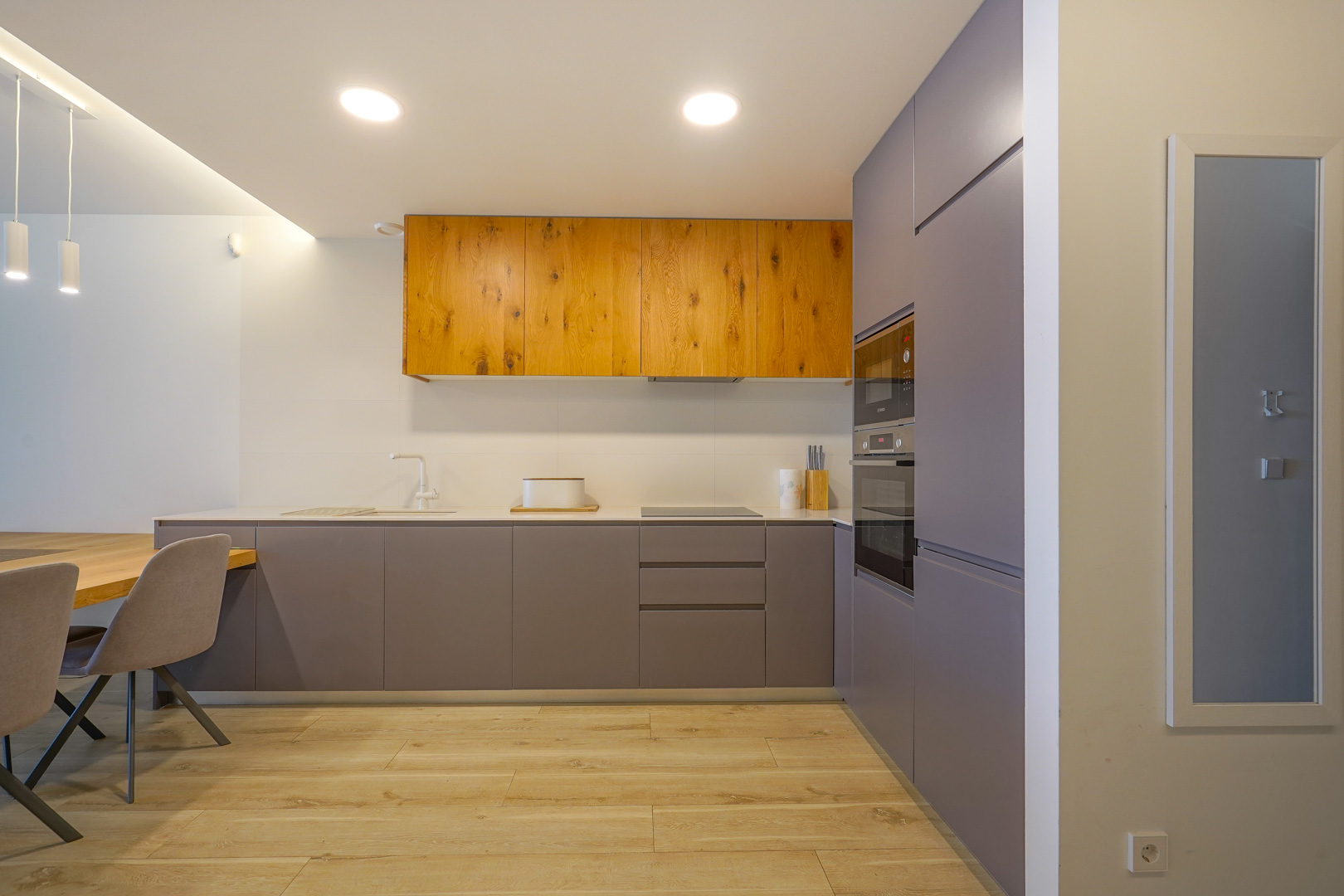 Moderne minimalistische keuken met matgrijze en natuurlijke houten kasten, voorzien van inbouwapparatuur, een wit werkblad en een lichte houtlook vloer.