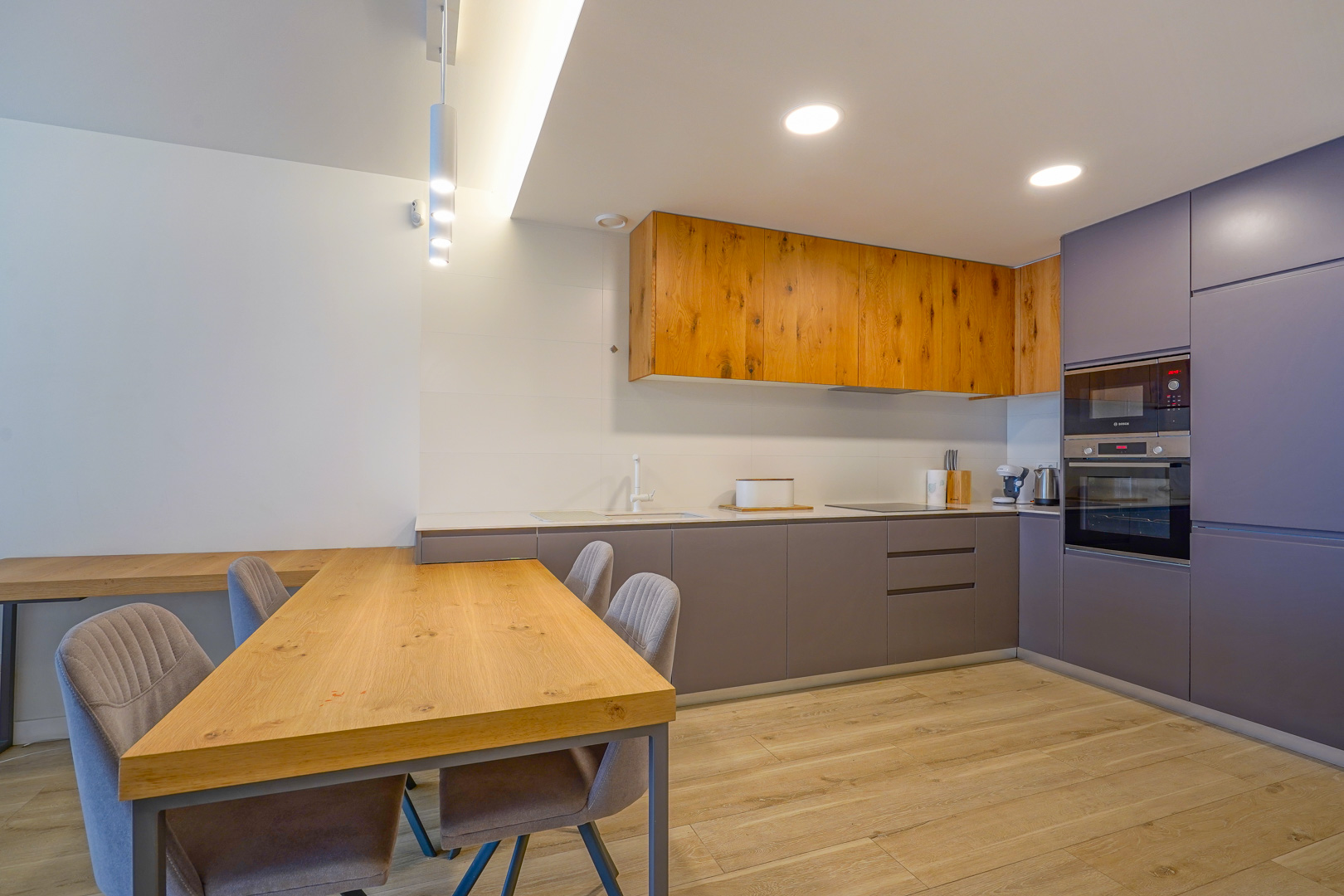 Moderne open keuken met kasten van natuurlijk hout en grijs, inbouwapparatuur en een bijpassende houten eettafel met gestoffeerde stoelen.