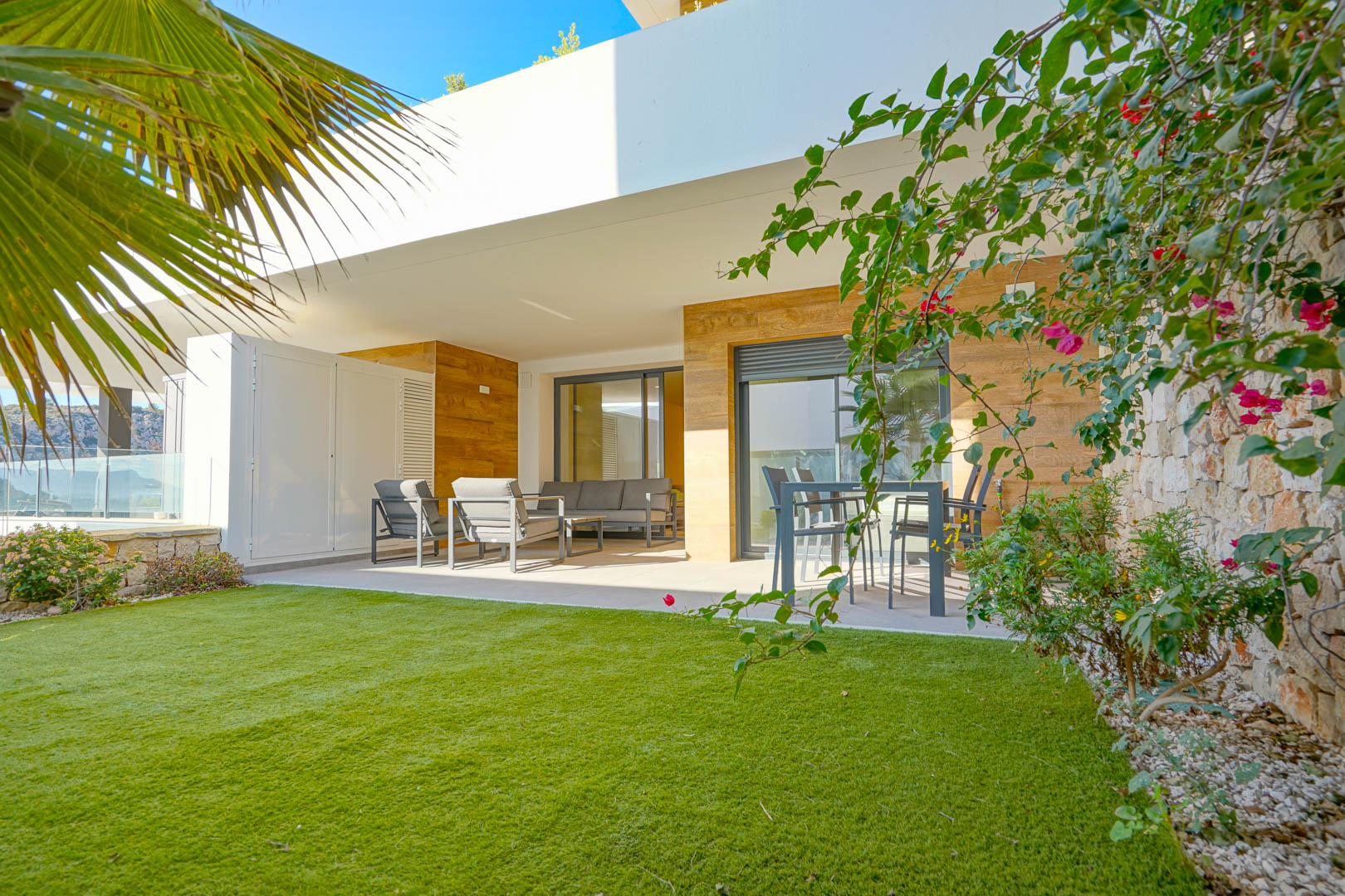 Overdekt terras met modern tuinmeubilair en een privétuin met kunstgras, omzoomd door een natuurstenen muur en mediterrane beplanting.