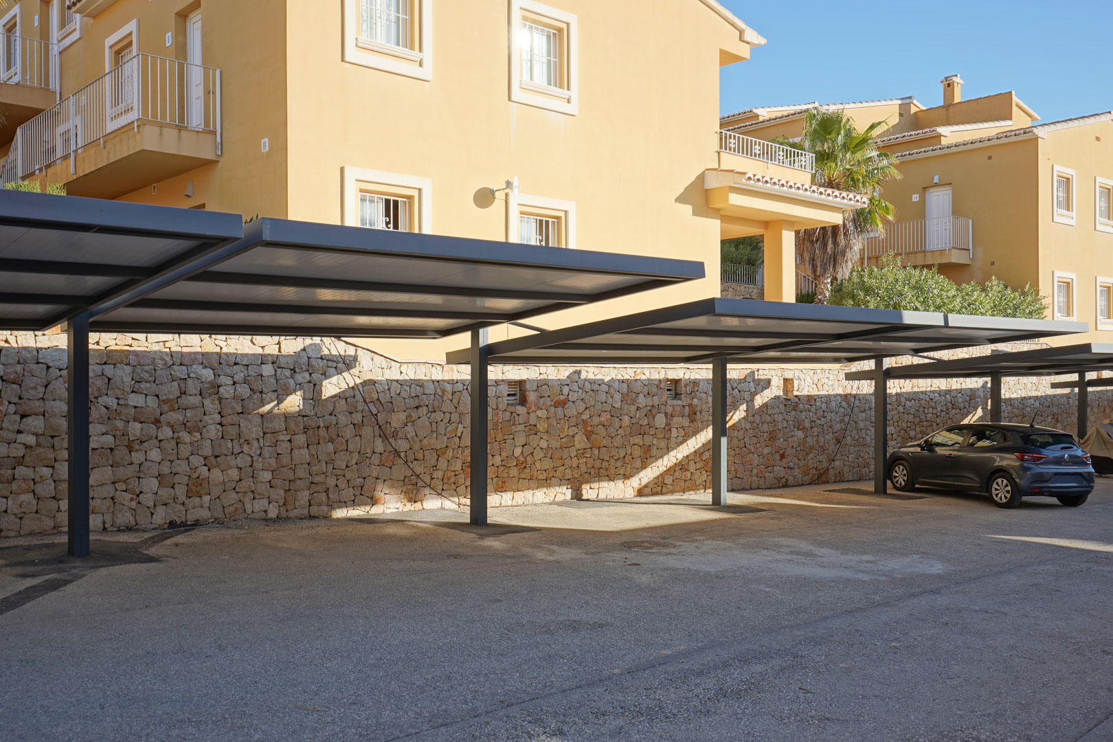 Overdekte parkeerplaats met moderne metalen carports en een natuurstenen muur, direct gelegen voor het appartementencomplex.