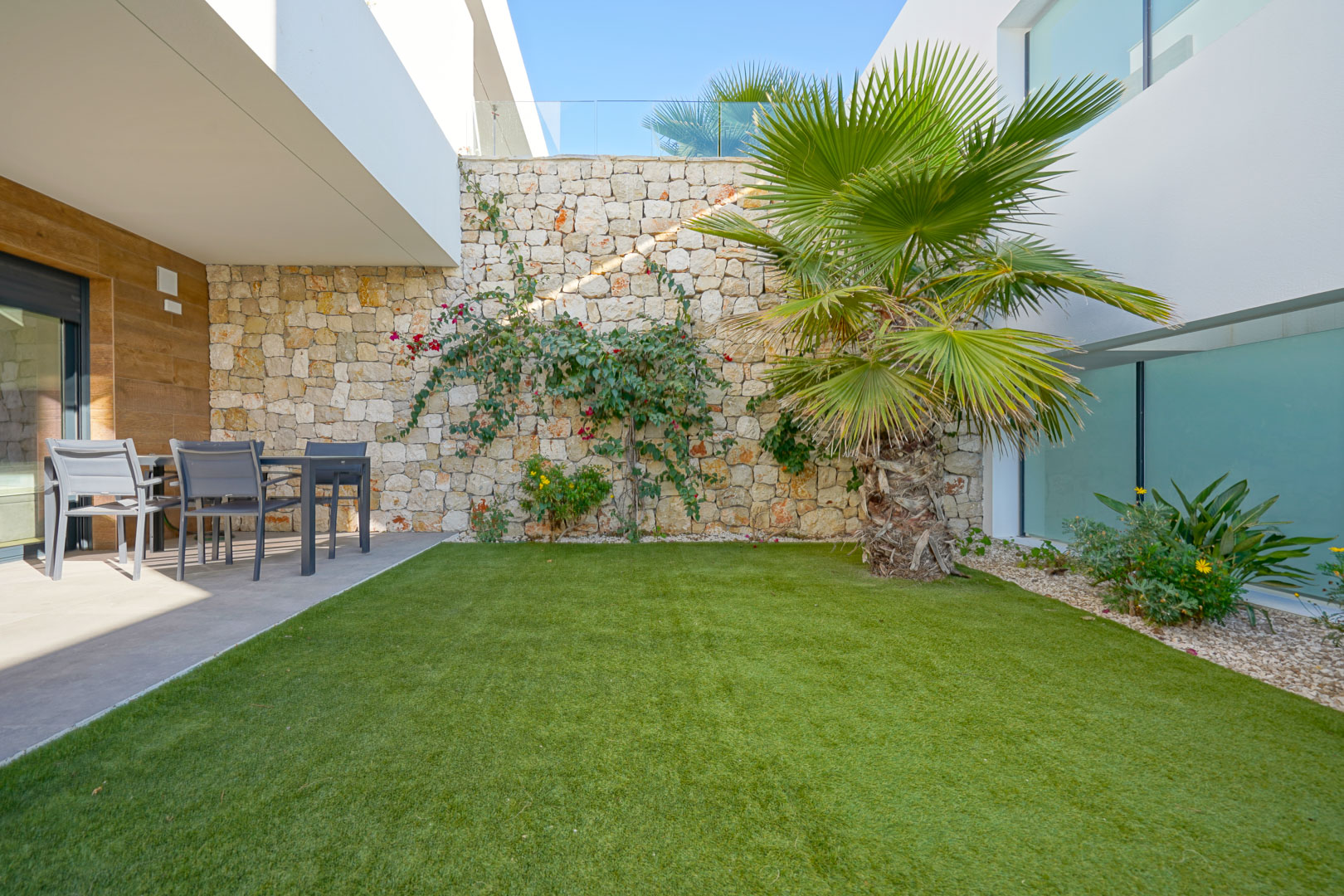 Privétuin met kunstgras, een palmboom en een natuurstenen muur. Een overdekt terras biedt een schaduwrijke eethoek in de buitenlucht met moderne afwerking.
