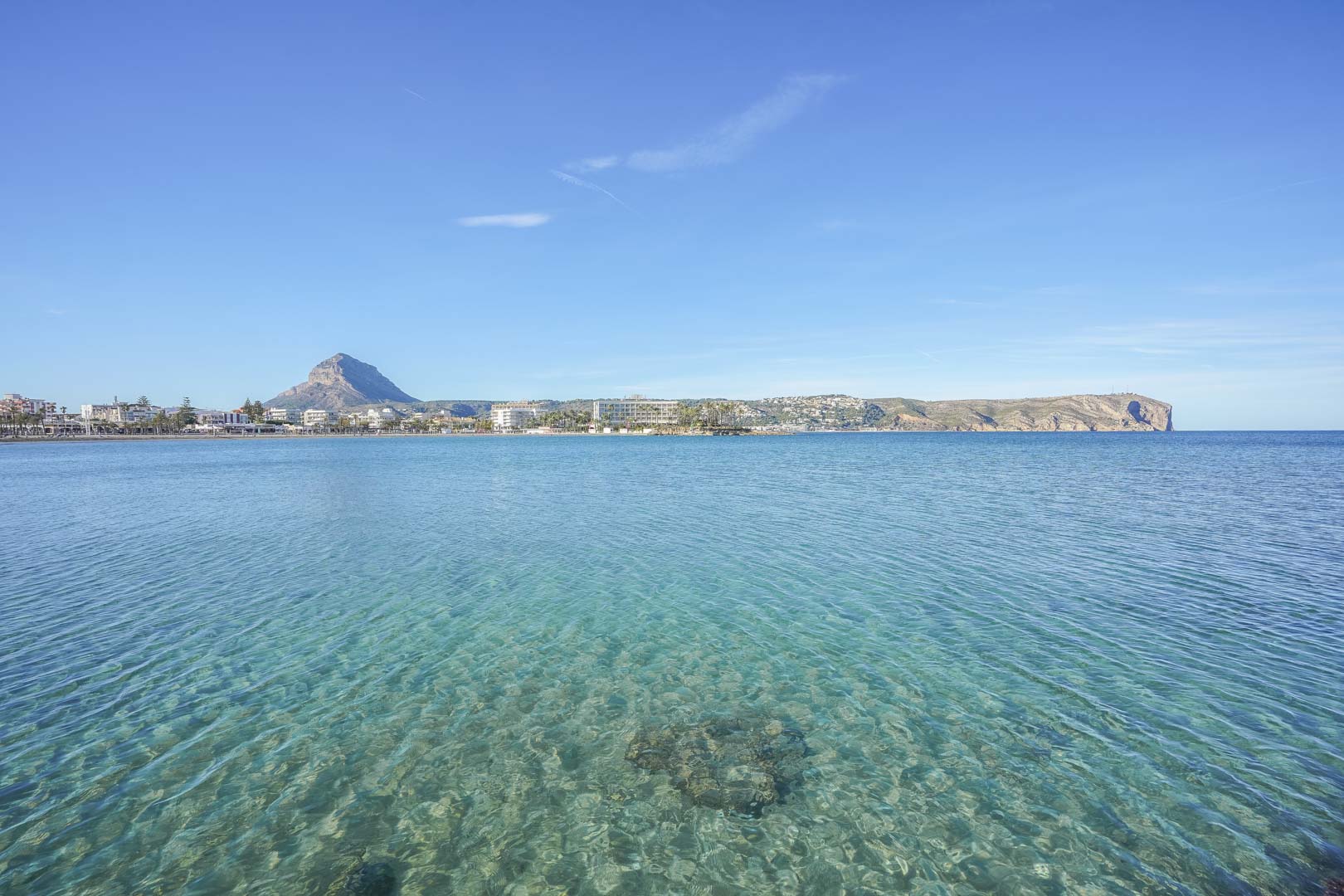 Panoramisch uitzicht op de baai van Jávea met kristalhelder turquoise water, de berg Montgó en de kliffen van Cabo de San Antonio aan de horizon.