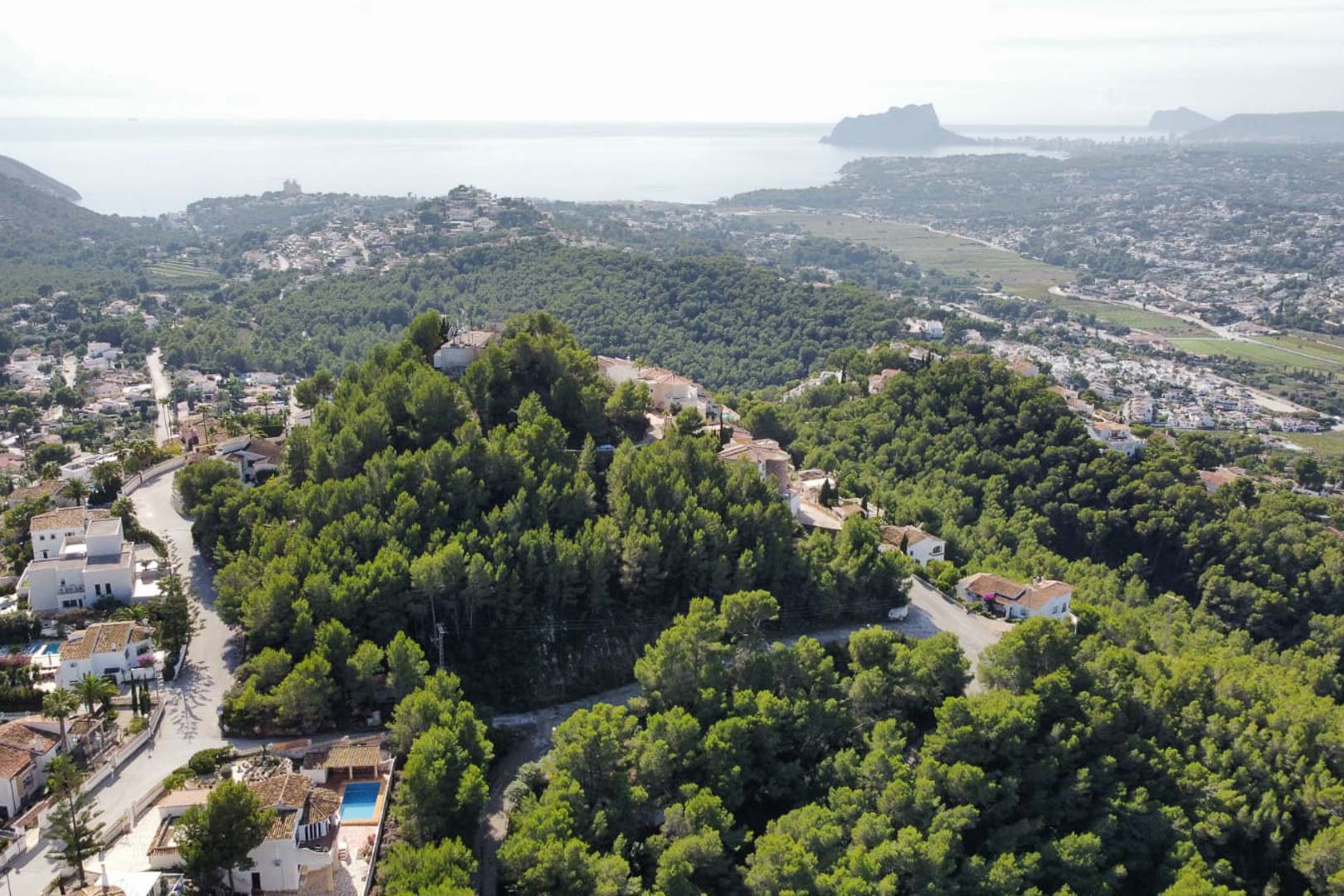 Luchtfoto van de woonwijk Paichi in Moraira, met dichte dennenbossen en een panoramisch uitzicht op de Middellandse Zee en de Peñón de Ifach.