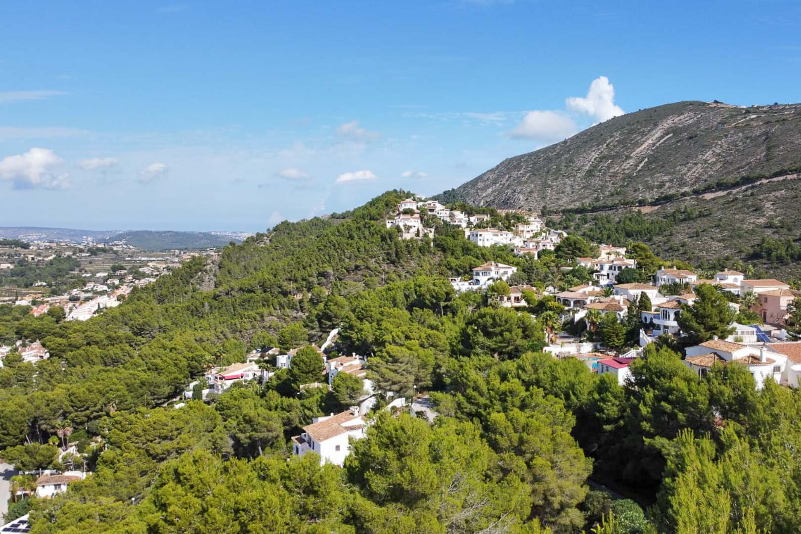 Luchtfoto van de woonwijk Paichi in Moraira, met witte villa's te midden van een dicht dennenbos en weids uitzicht op de omliggende bergen.