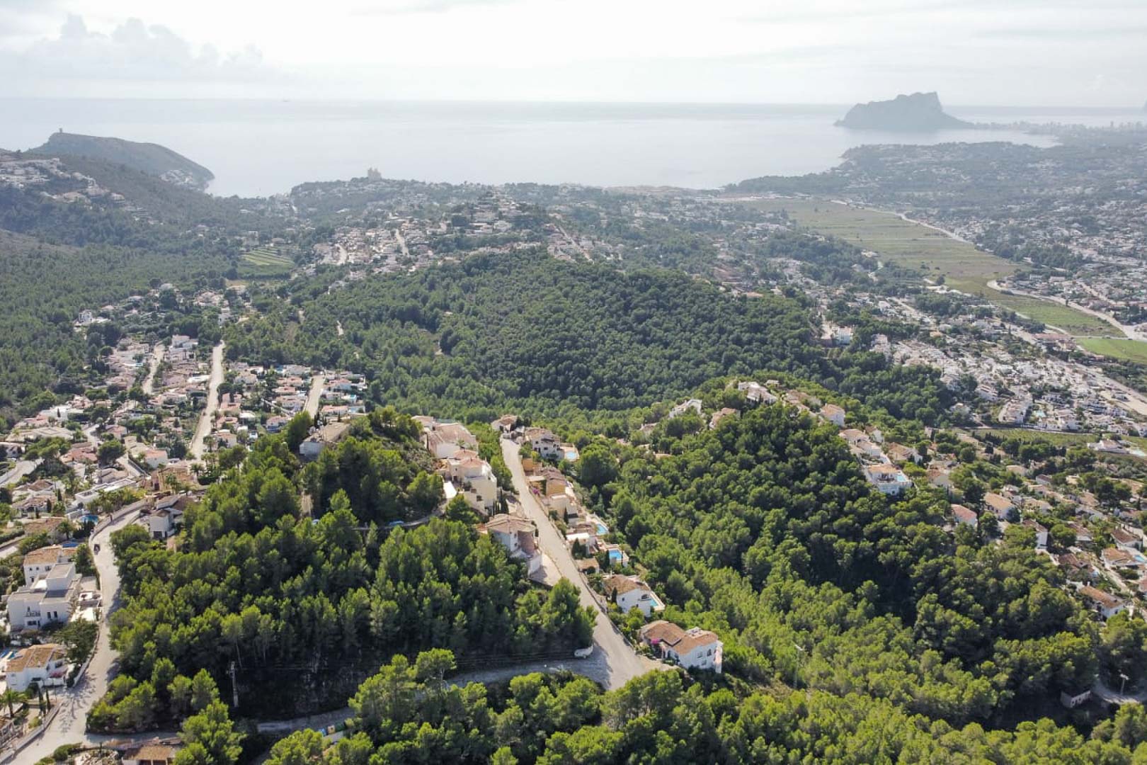 Panoramisch luchtfoto van de woonwijk Paichi, met dichte dennenbossen, vrijstaande villa's en de Peñón de Ifach aan de horizon van de Middellandse Zee.