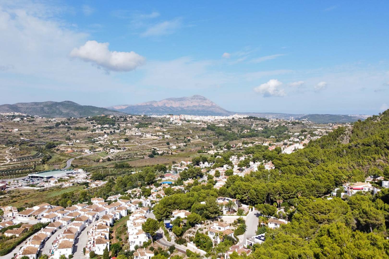 Verhoogd panoramisch uitzicht op mediterrane dennenbossen en witte villa's, met een opvallend bergsilhouet aan de horizon onder een heldere blauwe hemel.