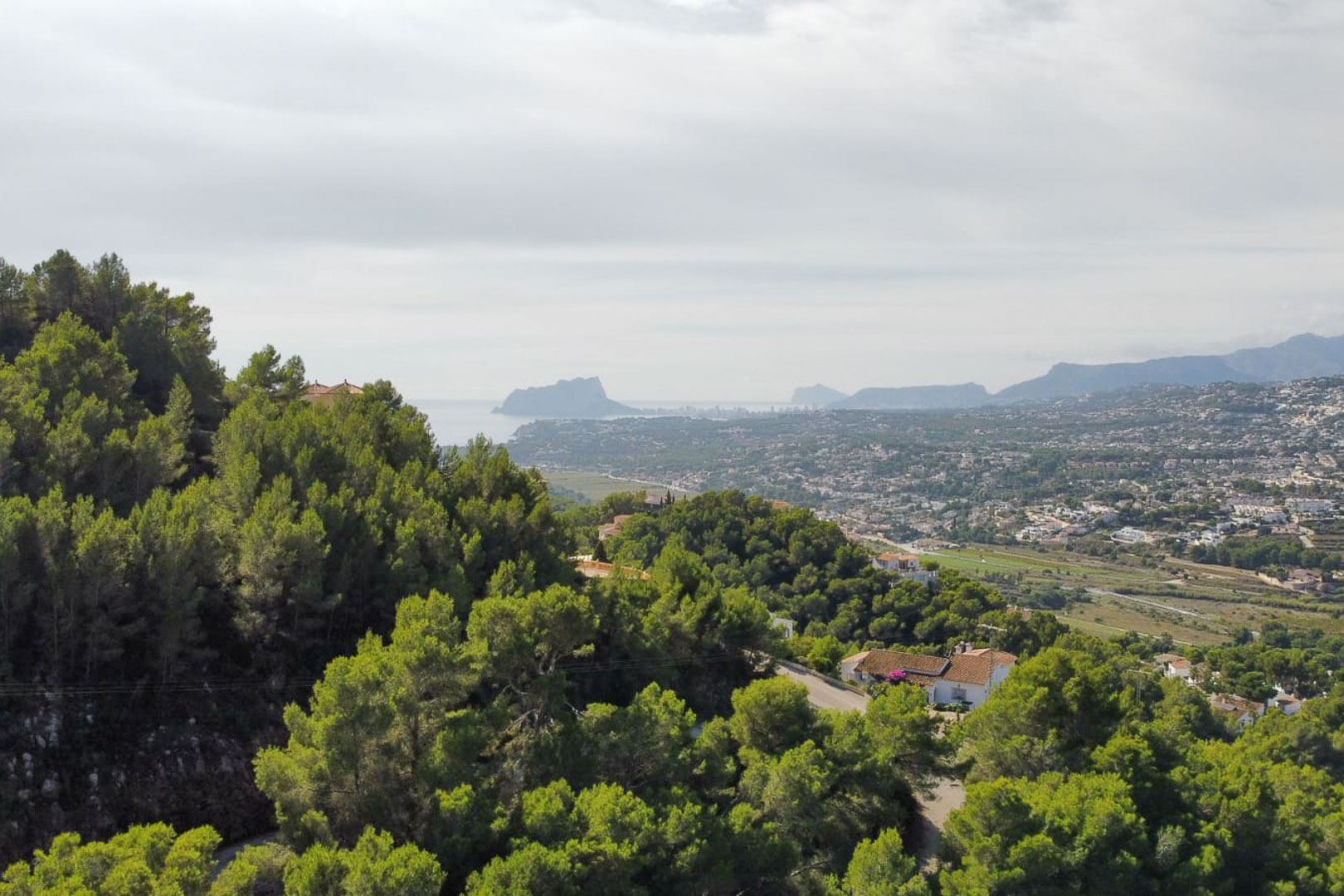Verhoogd panoramisch uitzicht vanaf het perceel in Paichi, met dichte dennenbossen, de residentiële vallei en het silhouet van de Peñón de Ifach aan de horizon.