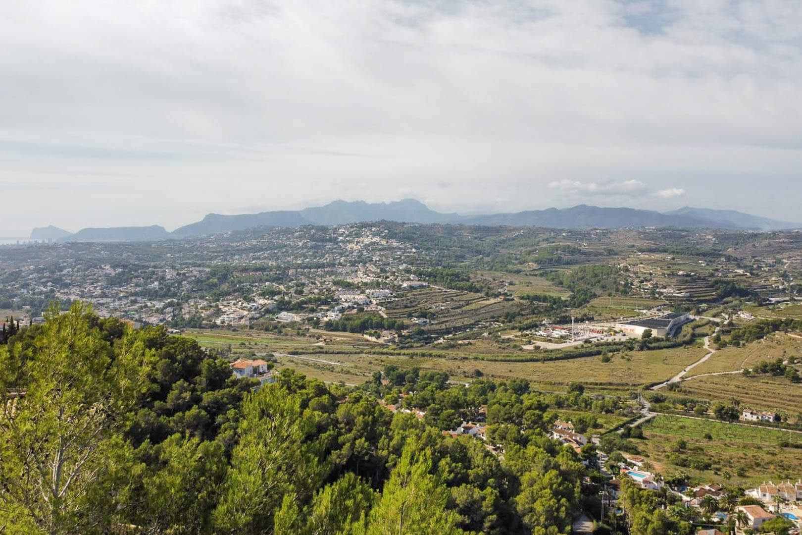 Verhoogd panoramisch uitzicht vanaf het perceel over de vallei van Moraira, met mediterrane dennenbossen en verre bergketens aan de horizon.