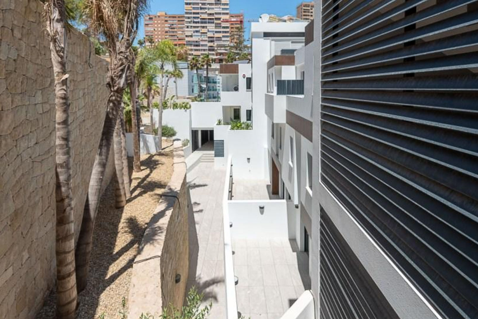 Buitenaanzicht van het complex met witte gevels, een natuurstenen muur en palmbomen. Moderne horizontale lamellen en geplaveide paden sieren de architectuur.